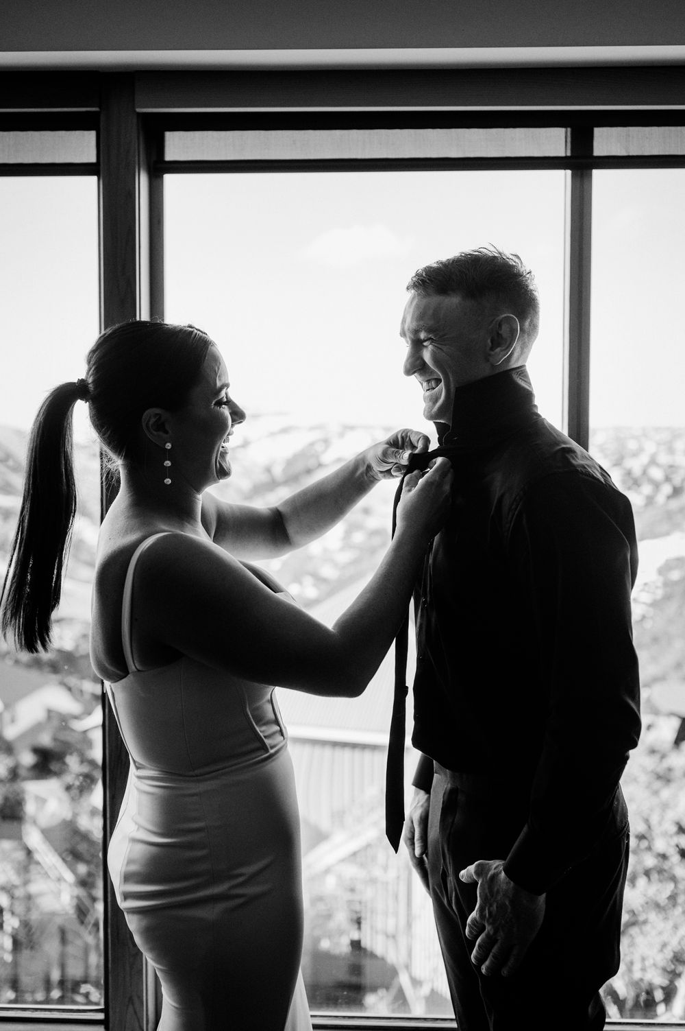A bride in a fitted dress adjusts the groom's tie in front of a window with scenic mountain views behind them.