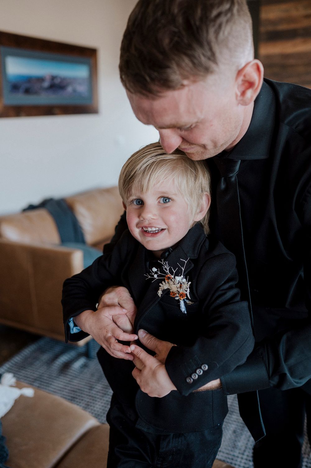 A tender moment of father and son sharing laughter while both wearing dark suits during a special occasion.