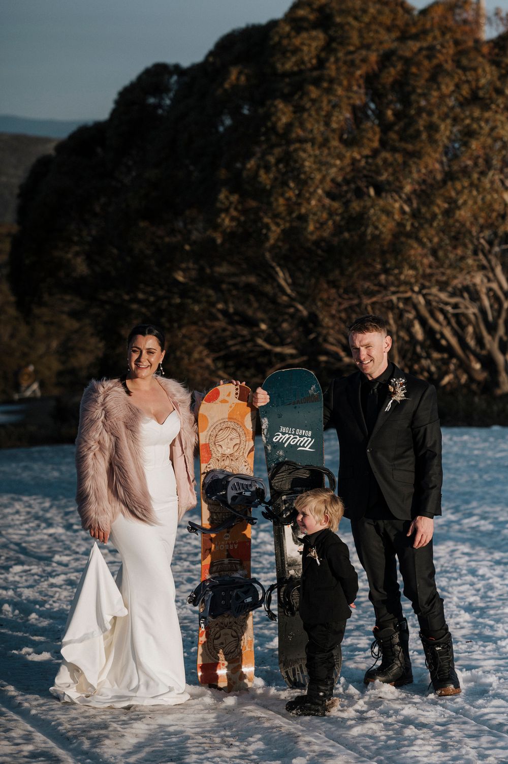 Family poses together in winter snow scene with mailbox, bride wearing white gown and fur jacket at outdoor wedding.