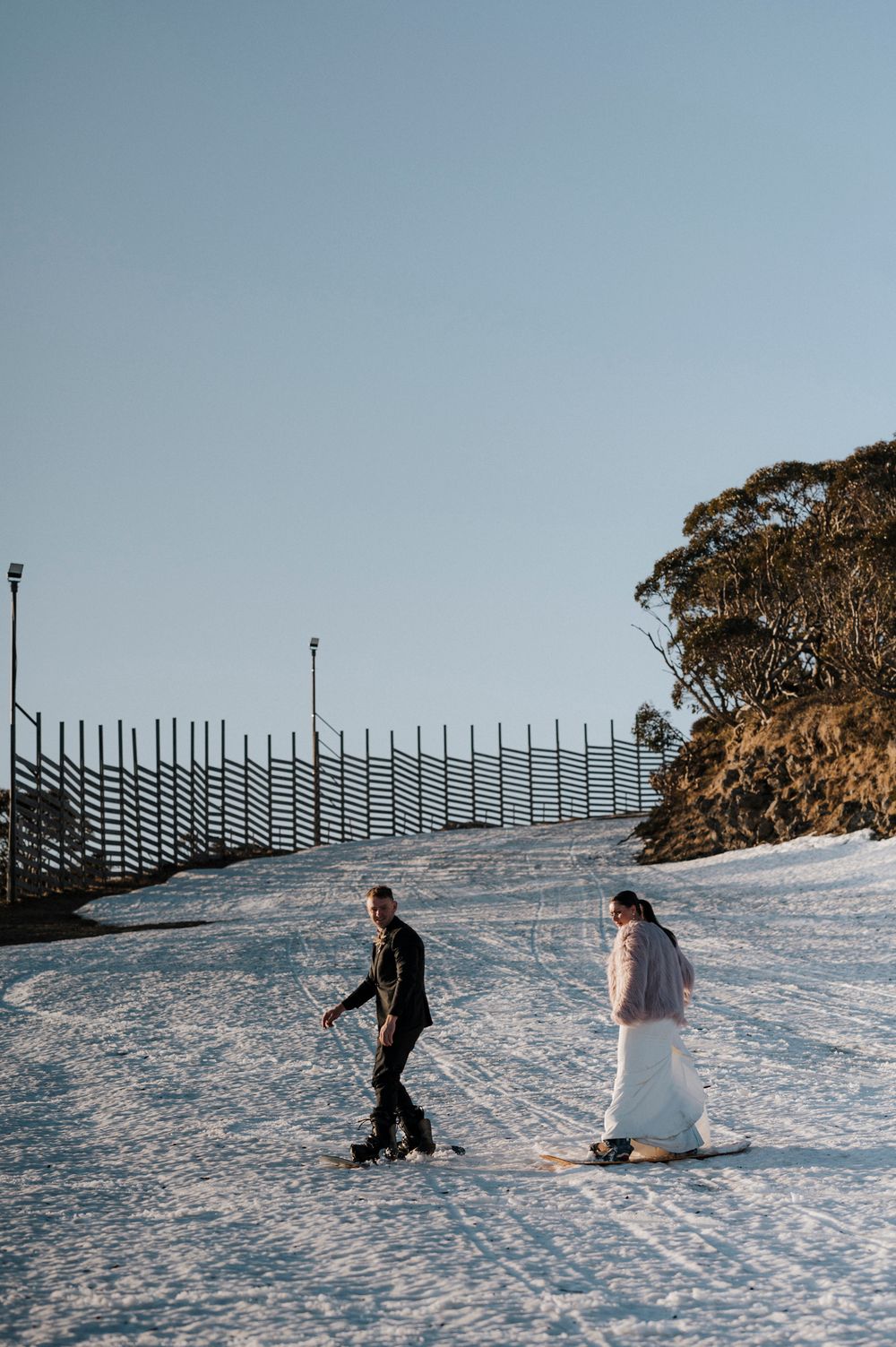 Two skiers making their way down a snowy slope with safety fencing and eucalyptus trees visible against a clear blue sky.