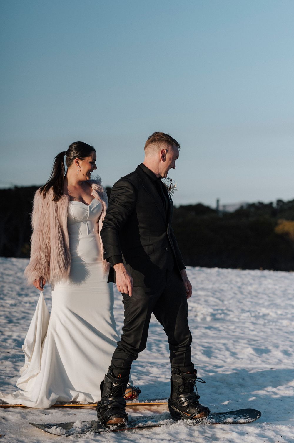 Wedding couple wearing winter attire poses together in deep snow during sunset photoshoot.