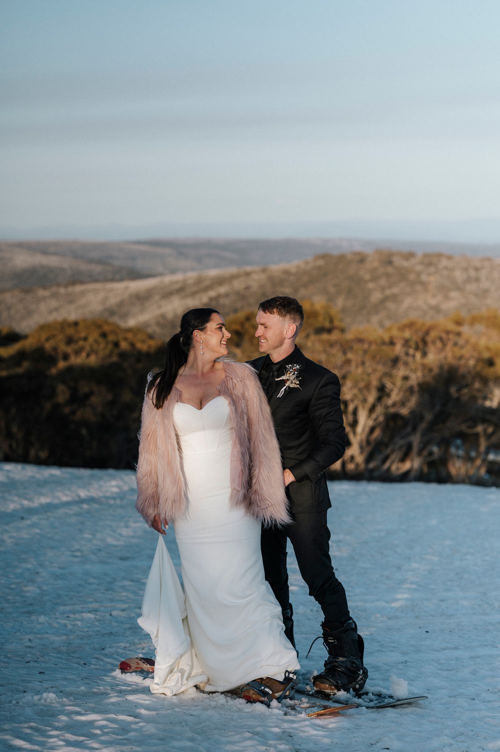 A couple poses in winter wedding attire on a snowy hilltop overlooking a dramatic mountain landscape.