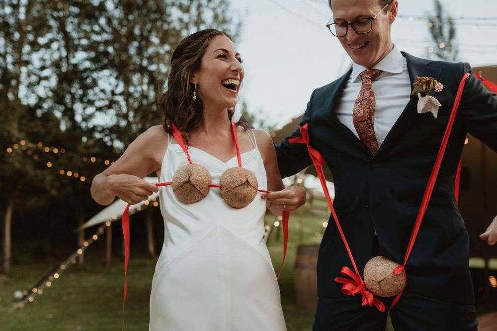 A joyful wedding couple sharing a lighthearted moment outdoors with string lights in the background.