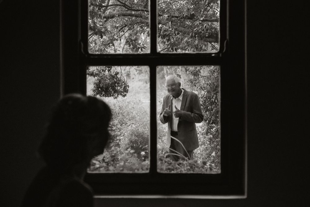Black and white image of father seeing his daughter, the bride through a window. He is giving her a thumbs up