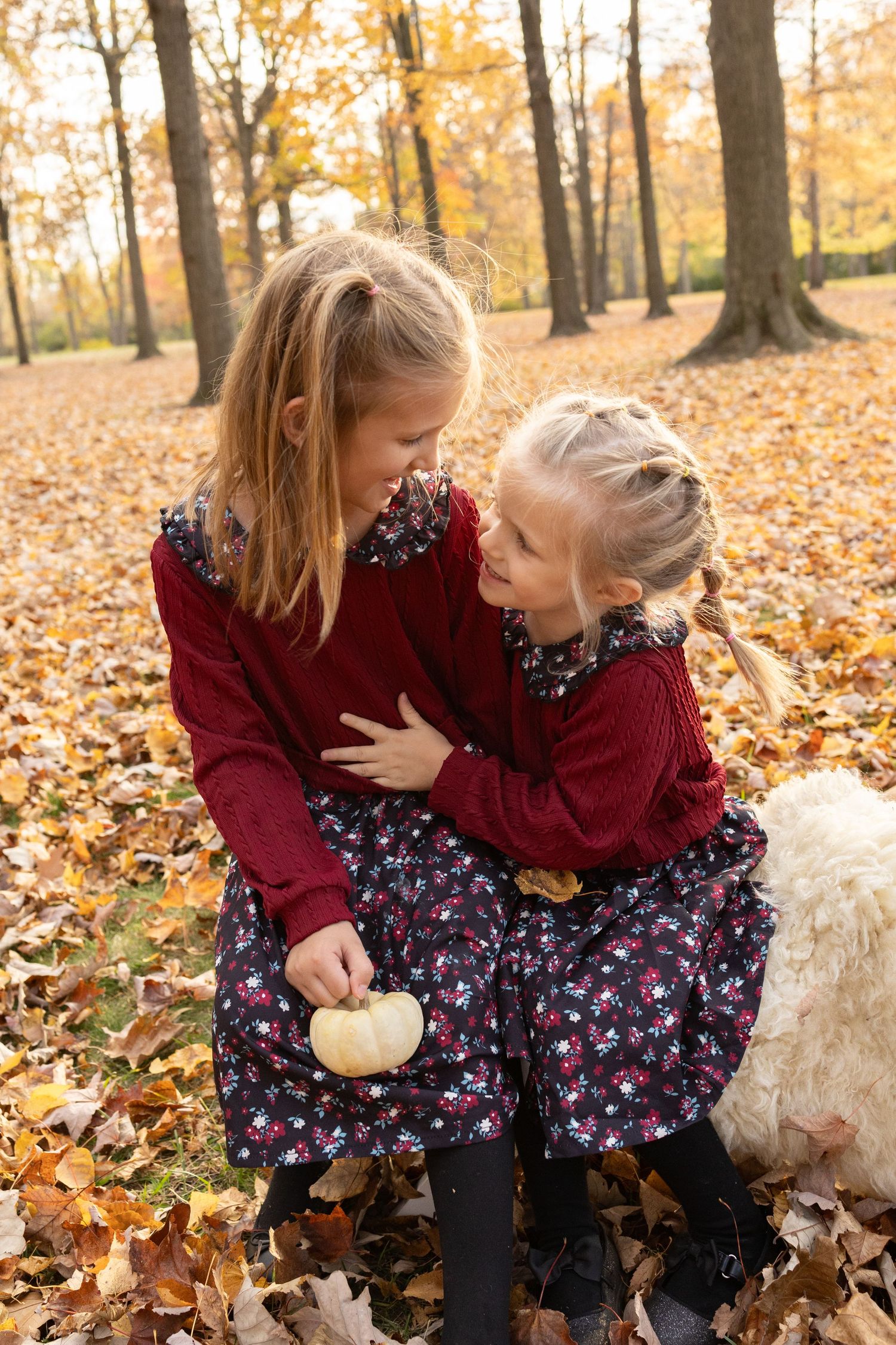 Two children in matching burgundy dresses share a moment among fallen autumn leaves.
