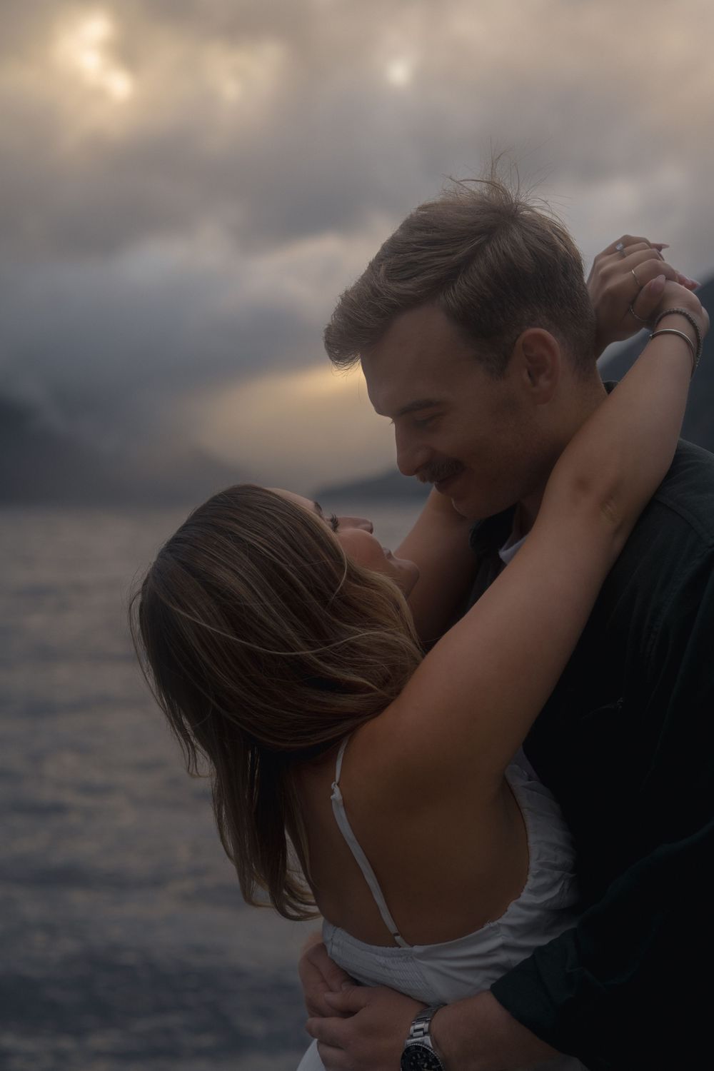 A romantic couple embraces against a moody sunset sky over water, creating an intimate silhouette.