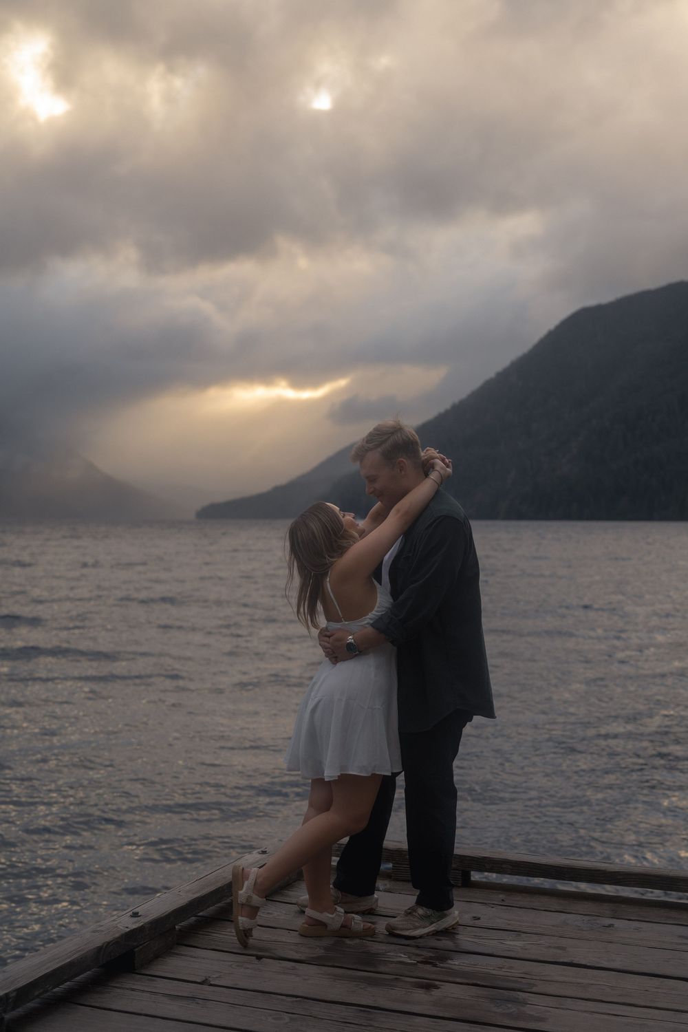 A romantic couple embraces on a wooden dock at sunset, with misty mountains and a lake creating a dramatic backdrop.