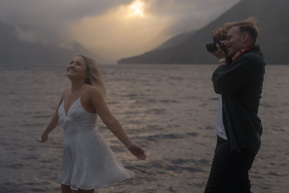 A couple embraces in silhouette against a dramatic sunset over a lake with mountains in the background.
