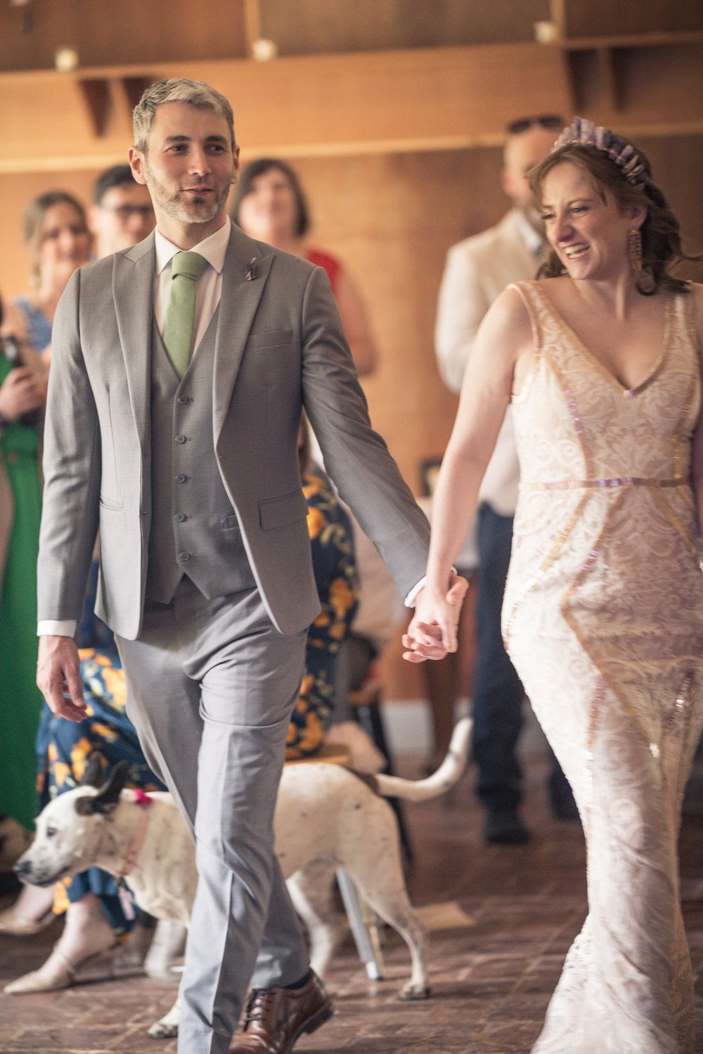 A couple in wedding attire walk hand in hand while guests and dogs watch in a rustic indoor setting.
