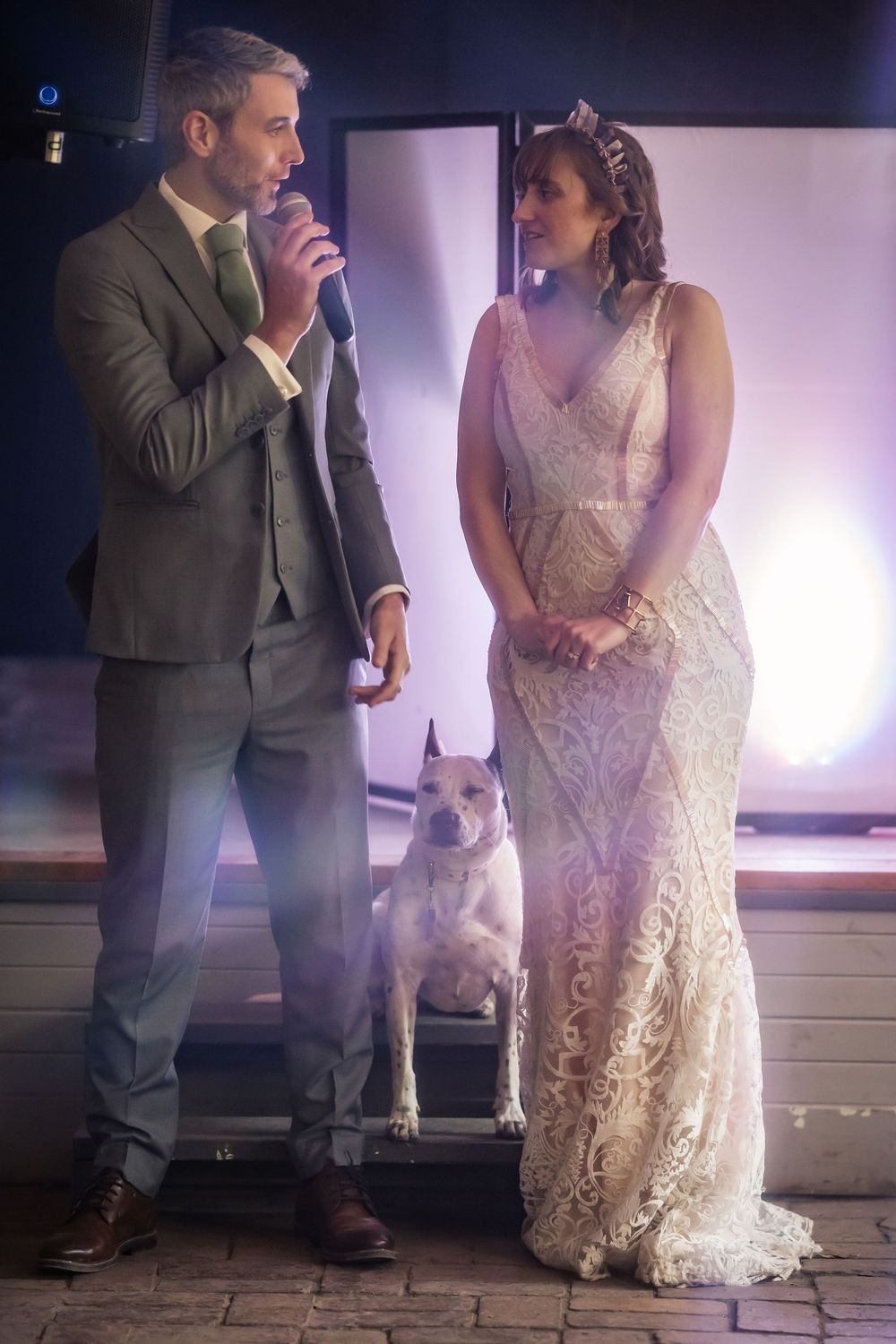A couple in wedding attire stands with their white dog during an indoor ceremony with soft pink lighting in the background.