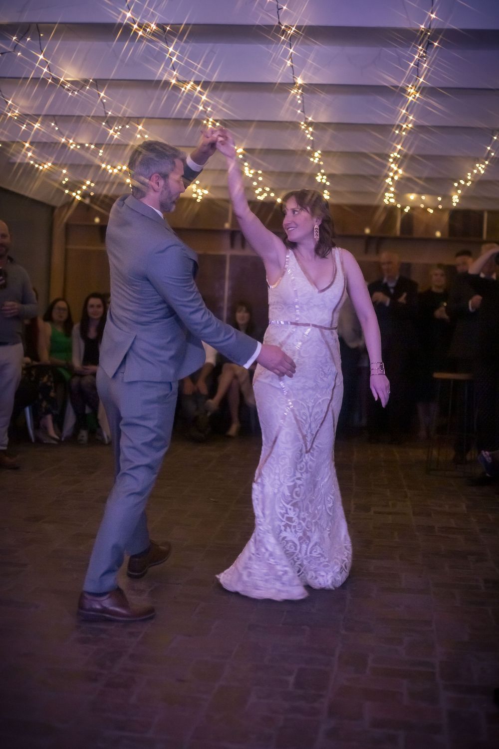 Couple dancing under twinkling string lights at a wedding reception with guests watching from the sides.