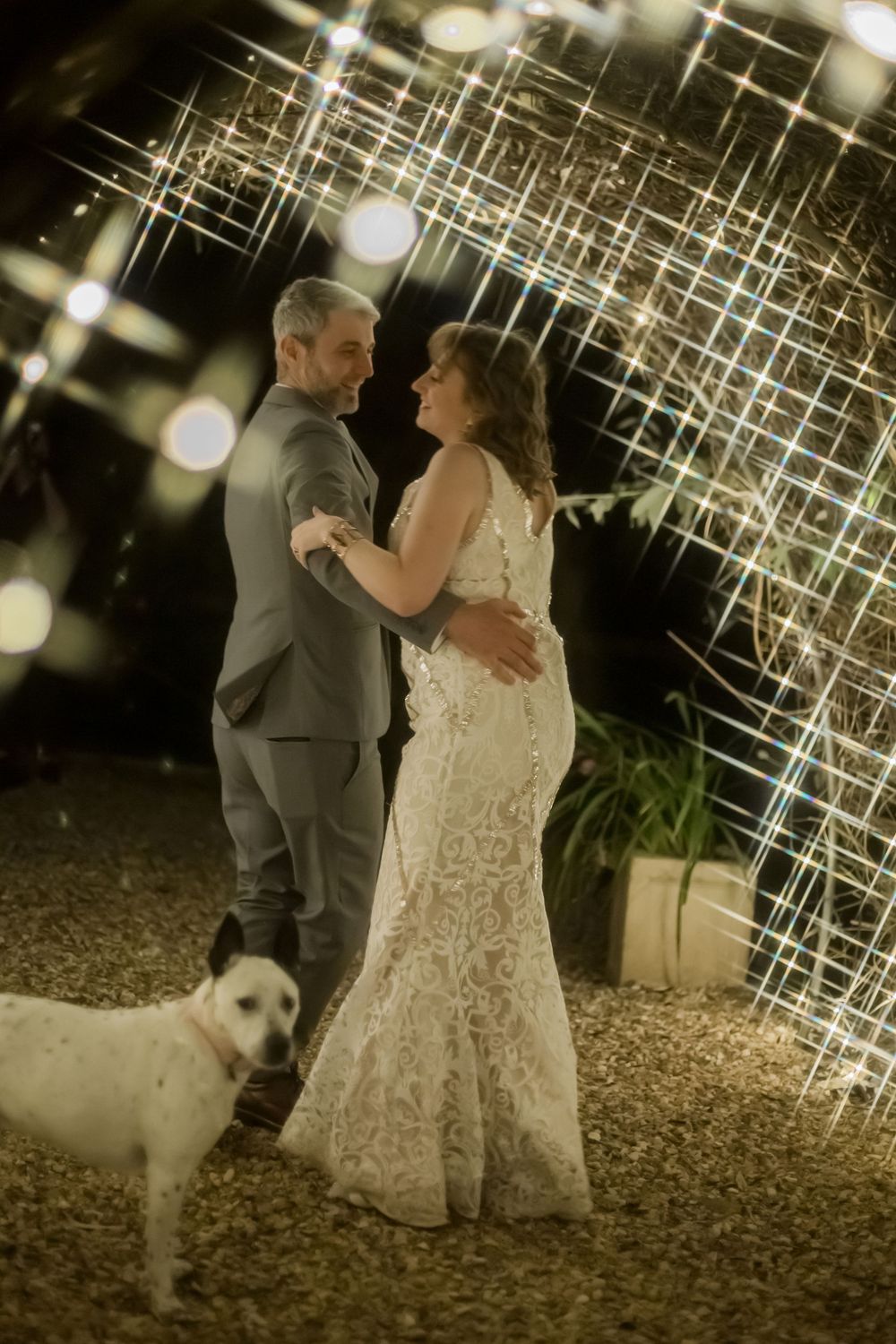 Couple sharing a romantic dance moment under twinkling string lights at a wedding reception with a white dog nearby.