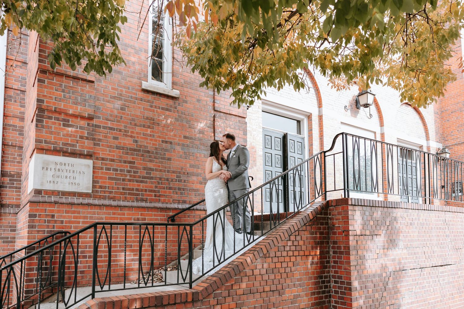 A couple stands on rustic brick staircase with wrought iron railing surrounded by autumn foliage outside historic building.