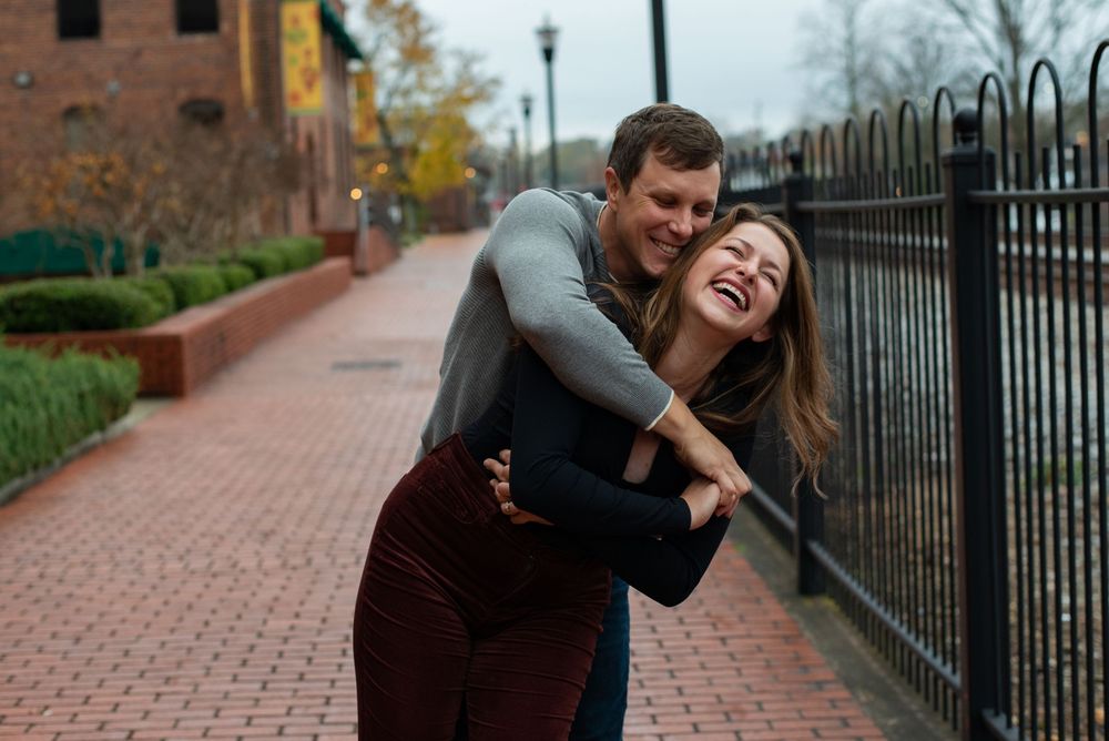 A couple shares a joyful, candid moment while embracing on a brick sidewalk beside a black iron fence.