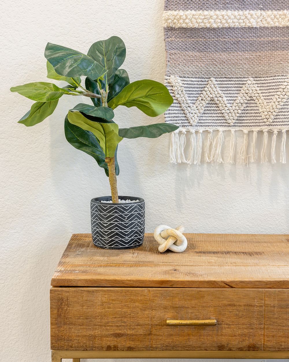 A fiddle leaf fig plant in a chevron-patterned pot sits on a wooden dresser with a decorative knot and woven wall art.
