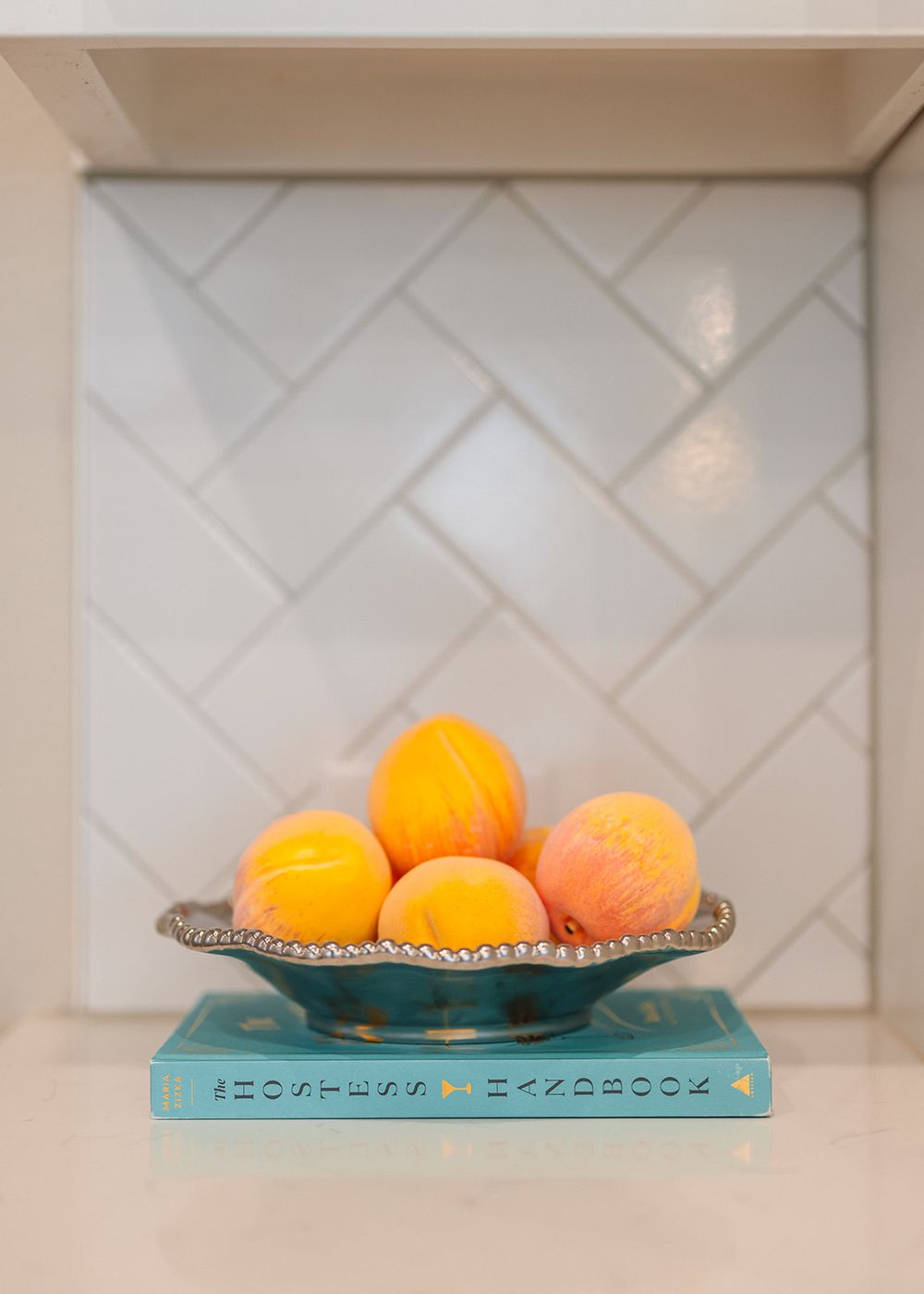 White herringbone tile backsplash behind glass shelf displaying bright orange citrus fruits in a teal serving dish.