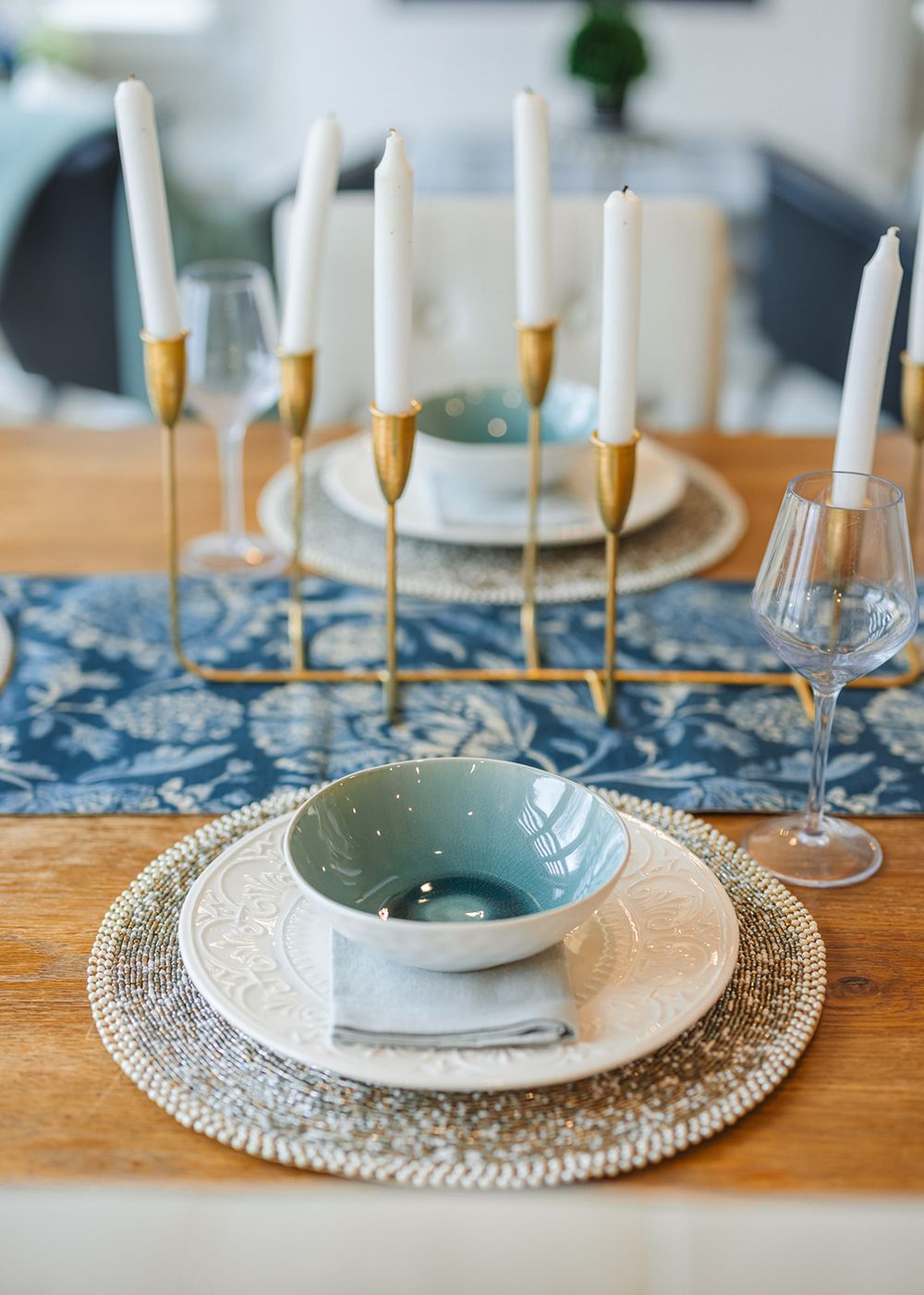 Elegant table setting with white candles in gold holders, woven placemats, blue runner and delicate dinnerware on wooden table.