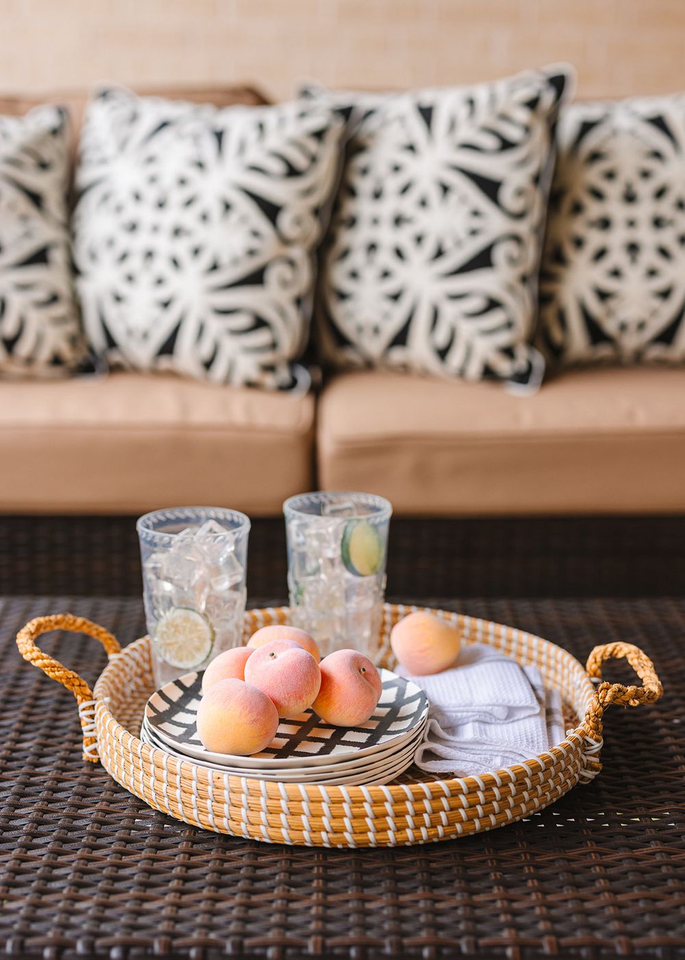 A woven tray with fresh peaches and glasses of water sits on a dark table in front of patterned throw pillows on a tan couch.