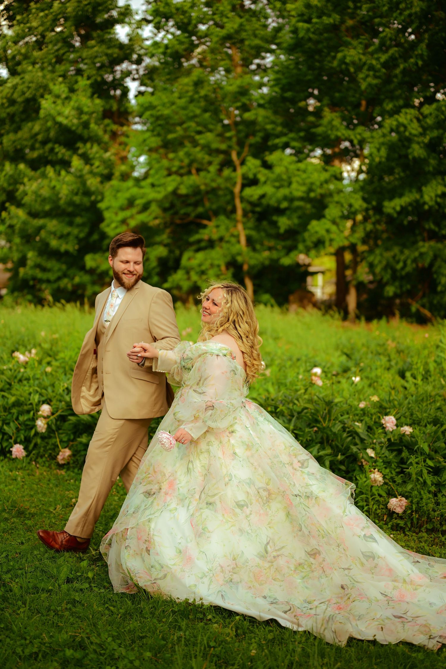 A couple in formalwear sits together in a lush green garden, the bride wearing a flowing floral patterned gown.