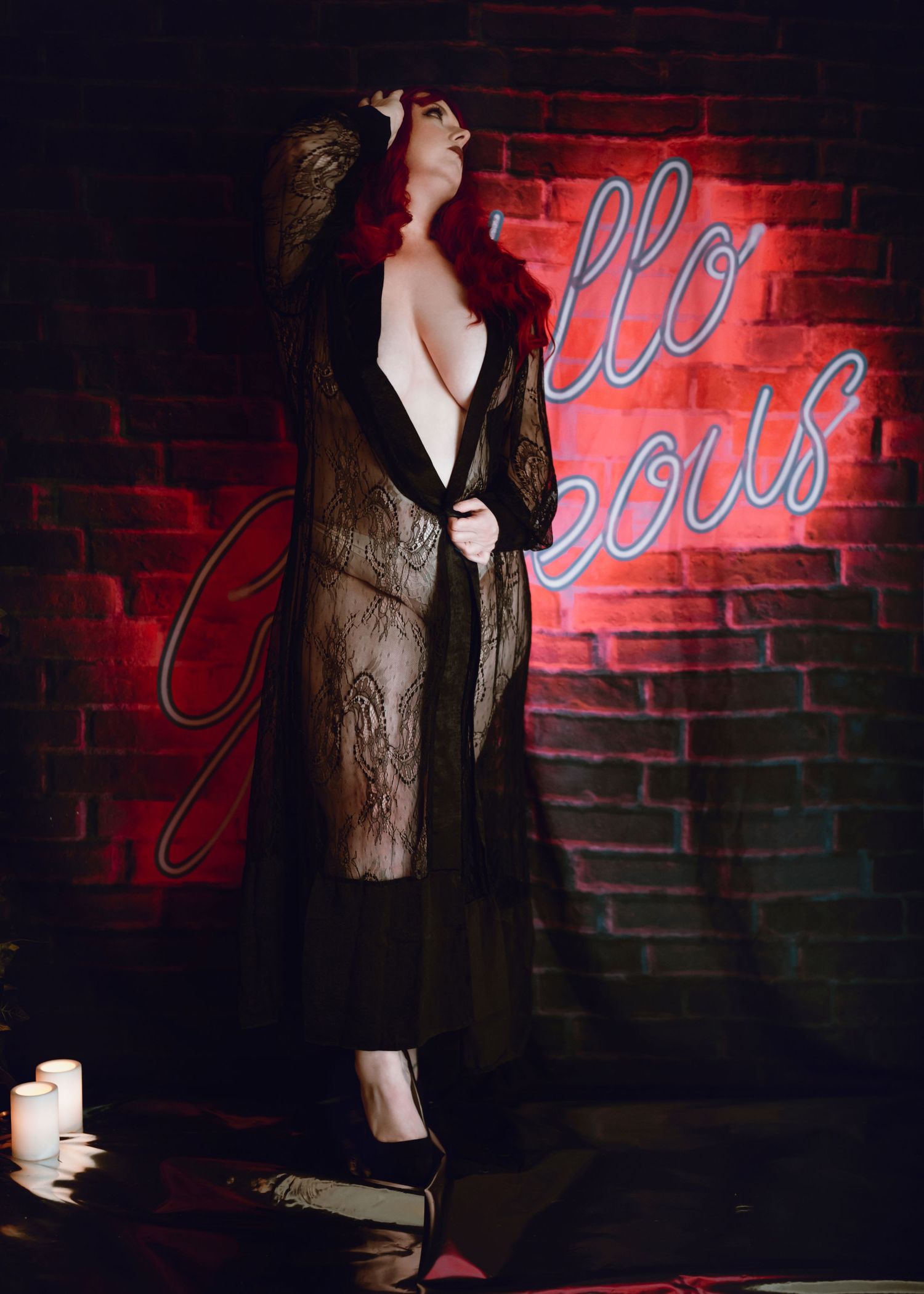 Fashion model in black dress and lace jacket poses against red brick wall with neon lighting creating dramatic atmosphere.