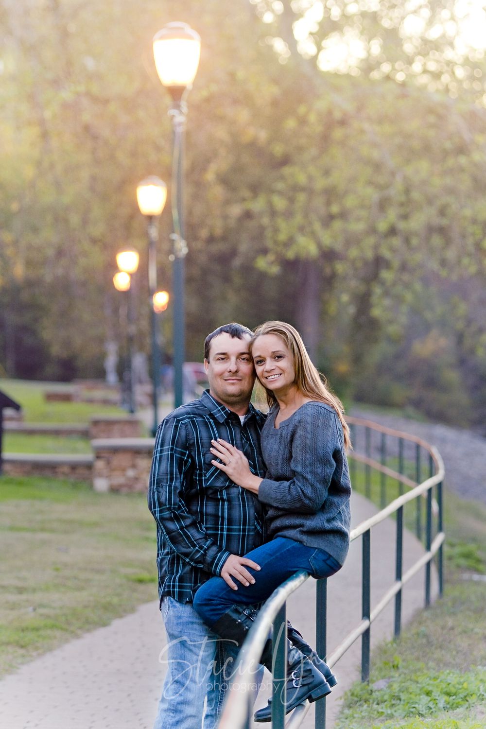 Couple poses together on path with glowing street lamps and greenery in evening light.