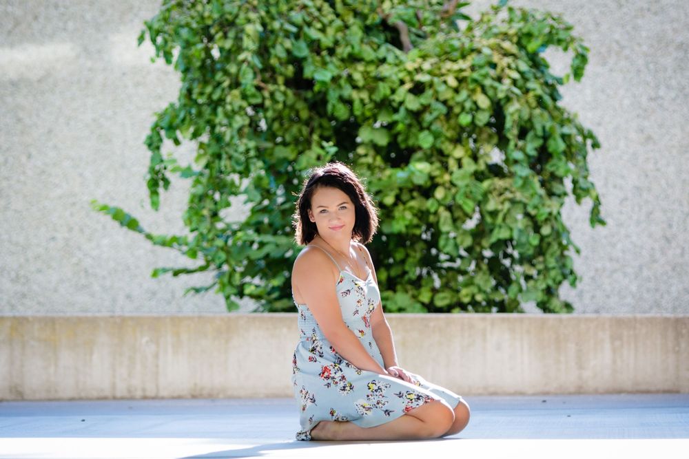 A figure in a floral dress sits on a concrete patio near lush green plants and foliage.