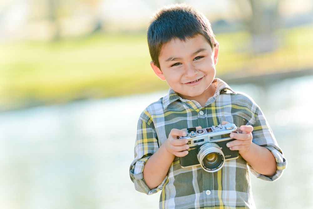 Child with a cheerful expression holding a vintage camera outdoors wearing a plaid shirt in bright sunlight.