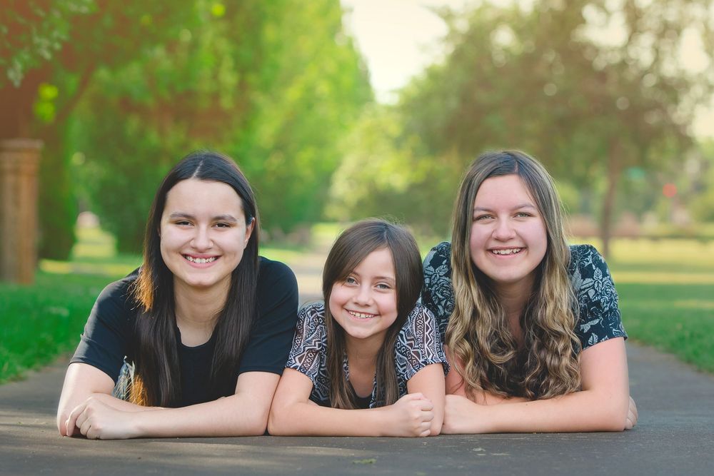 Three people lying together on grass outdoors smiling at the camera during a sunny day.