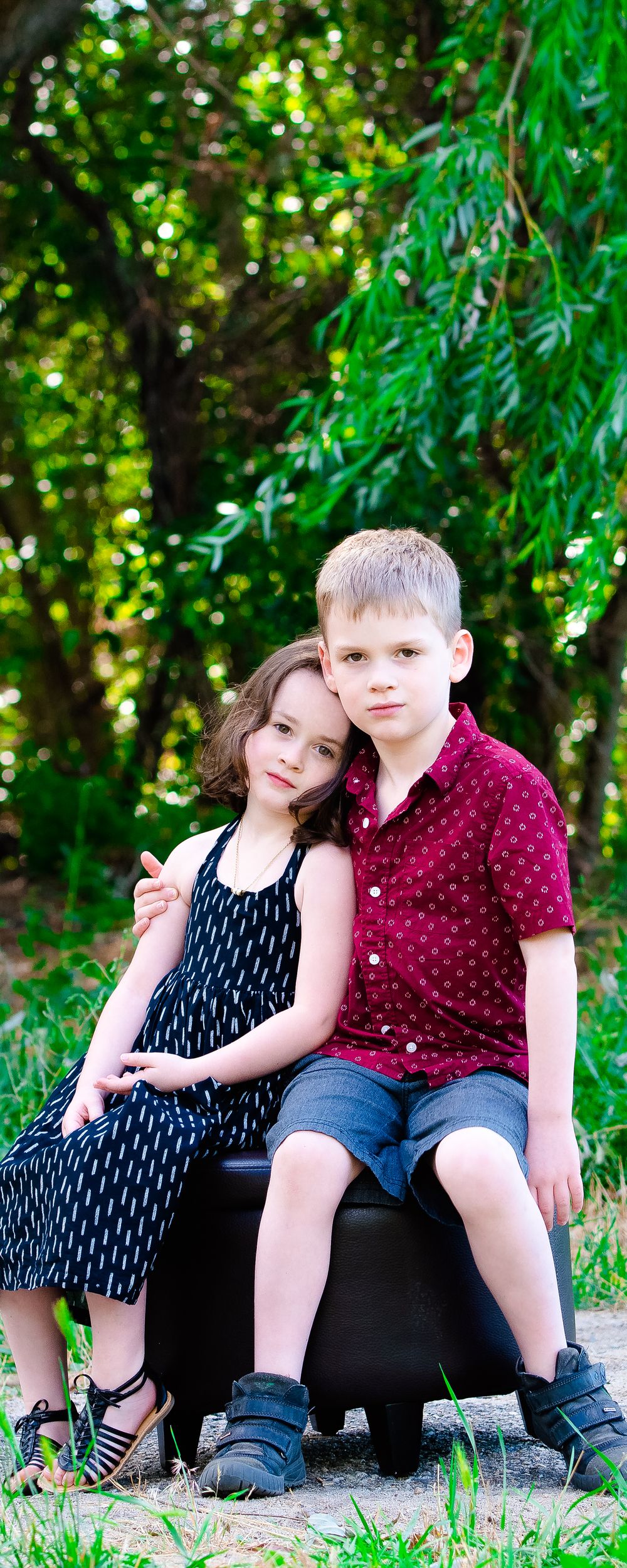 Two children sit close together on a black bench outdoors with lush green foliage in the background.