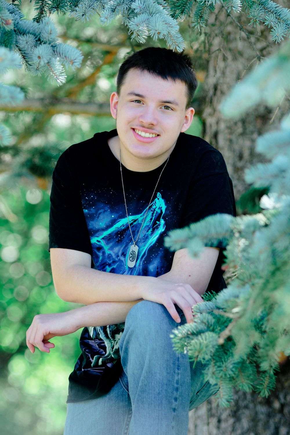 A person in a black t-shirt with blue design sits casually among evergreen branches in an outdoor portrait setting.
