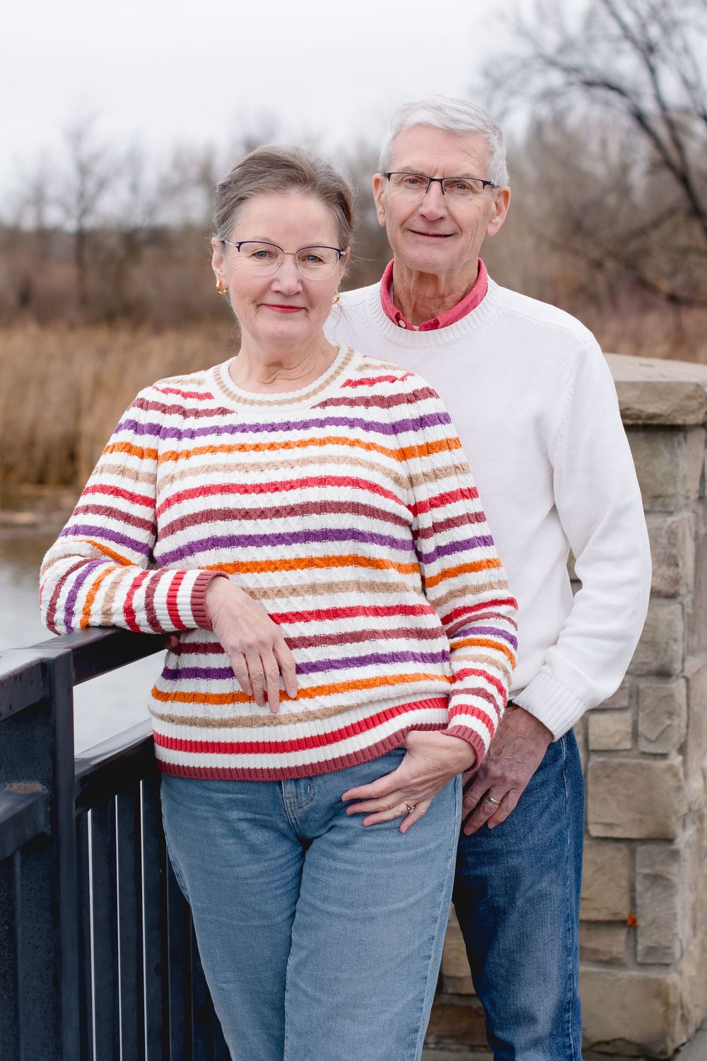 Elderly couple poses together by stone wall while wearing matching red and white striped sweaters and jeans.