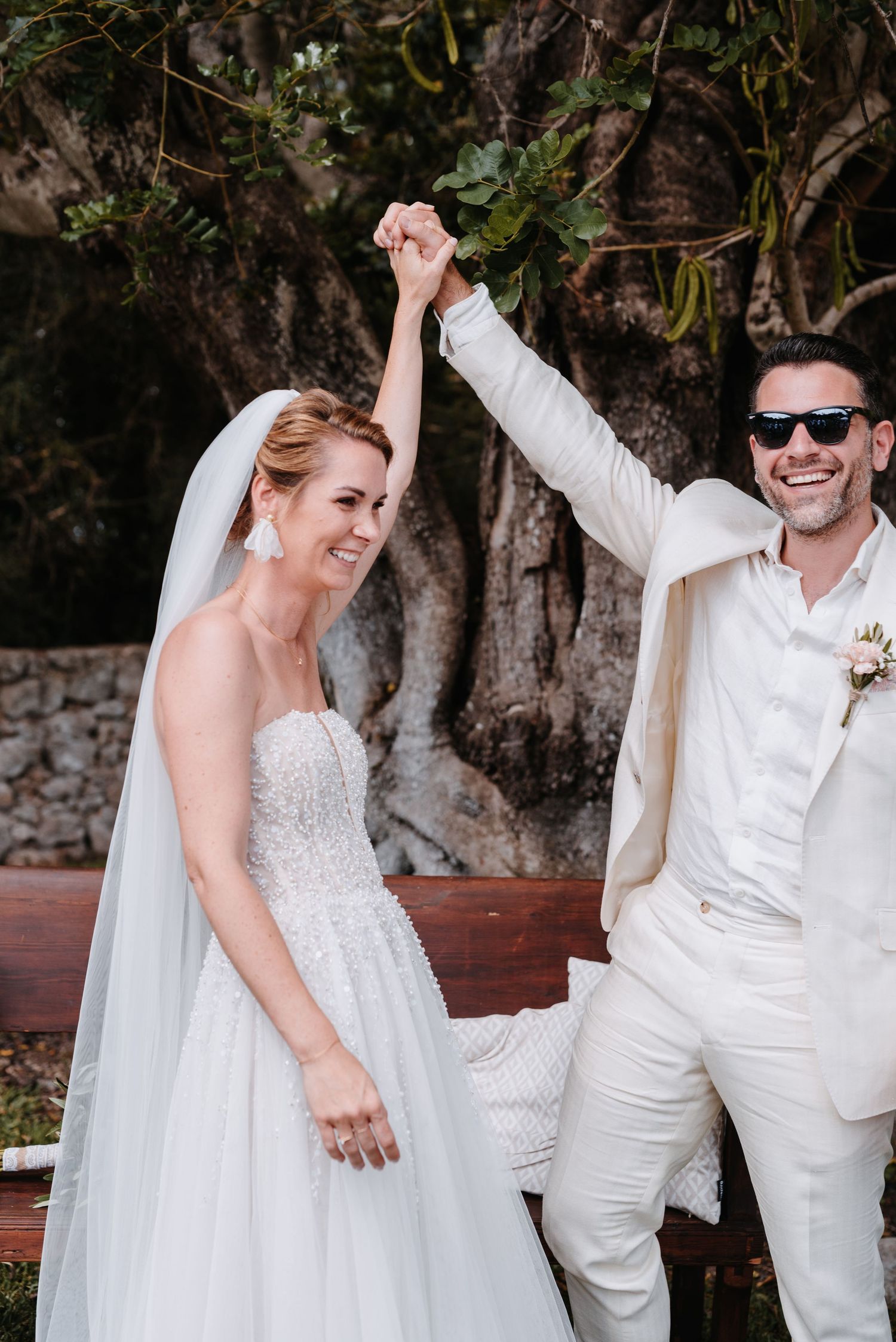 Das Hochzeitspaar feiert fröhlich in einem Garten im Freien, wobei die Braut ein weißes Kleid und der Bräutigam einen hellen Anzug trägt.