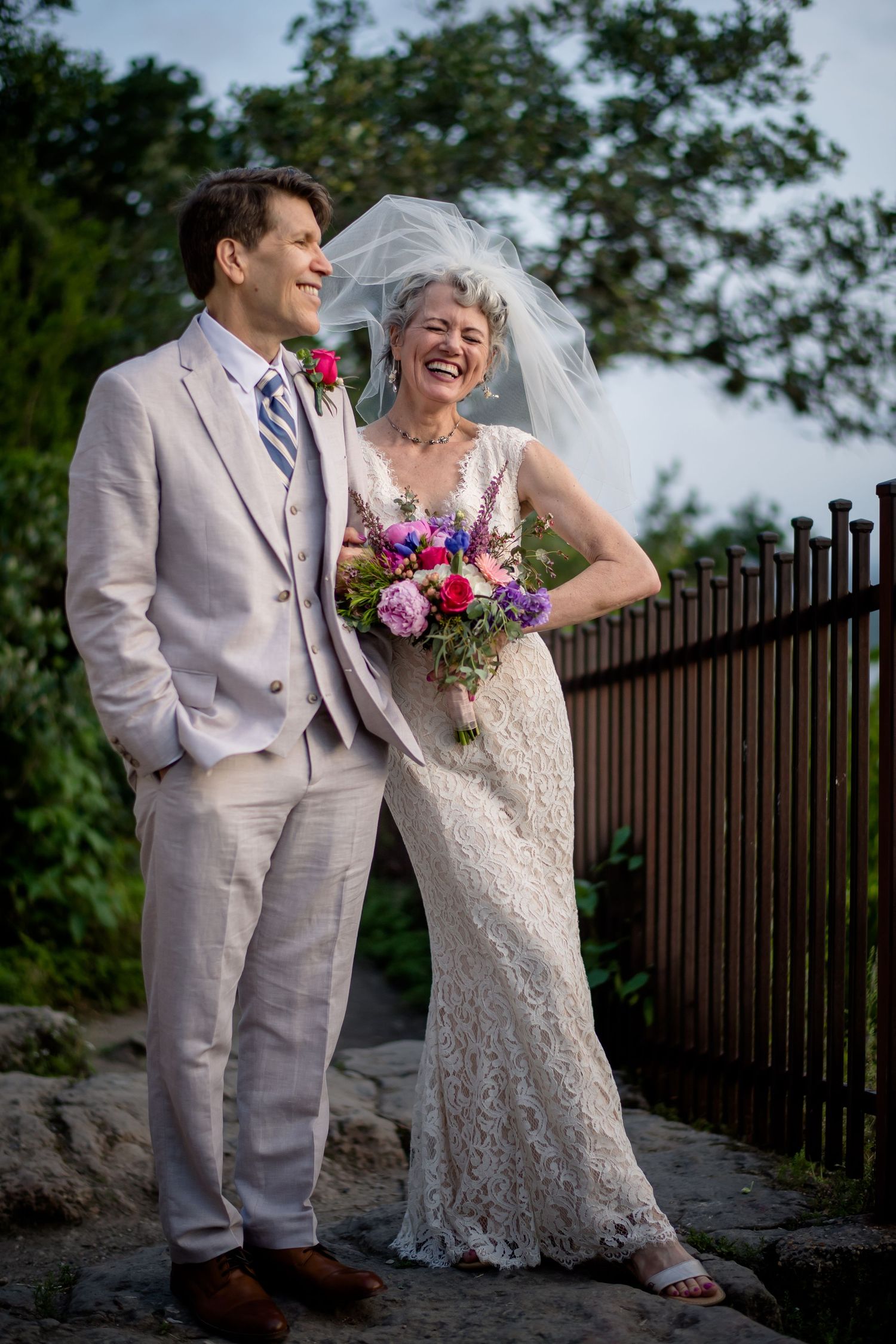Newlywed couple shares a happy moment outdoors, with bride in lace dress holding colorful bouquet by iron fence.