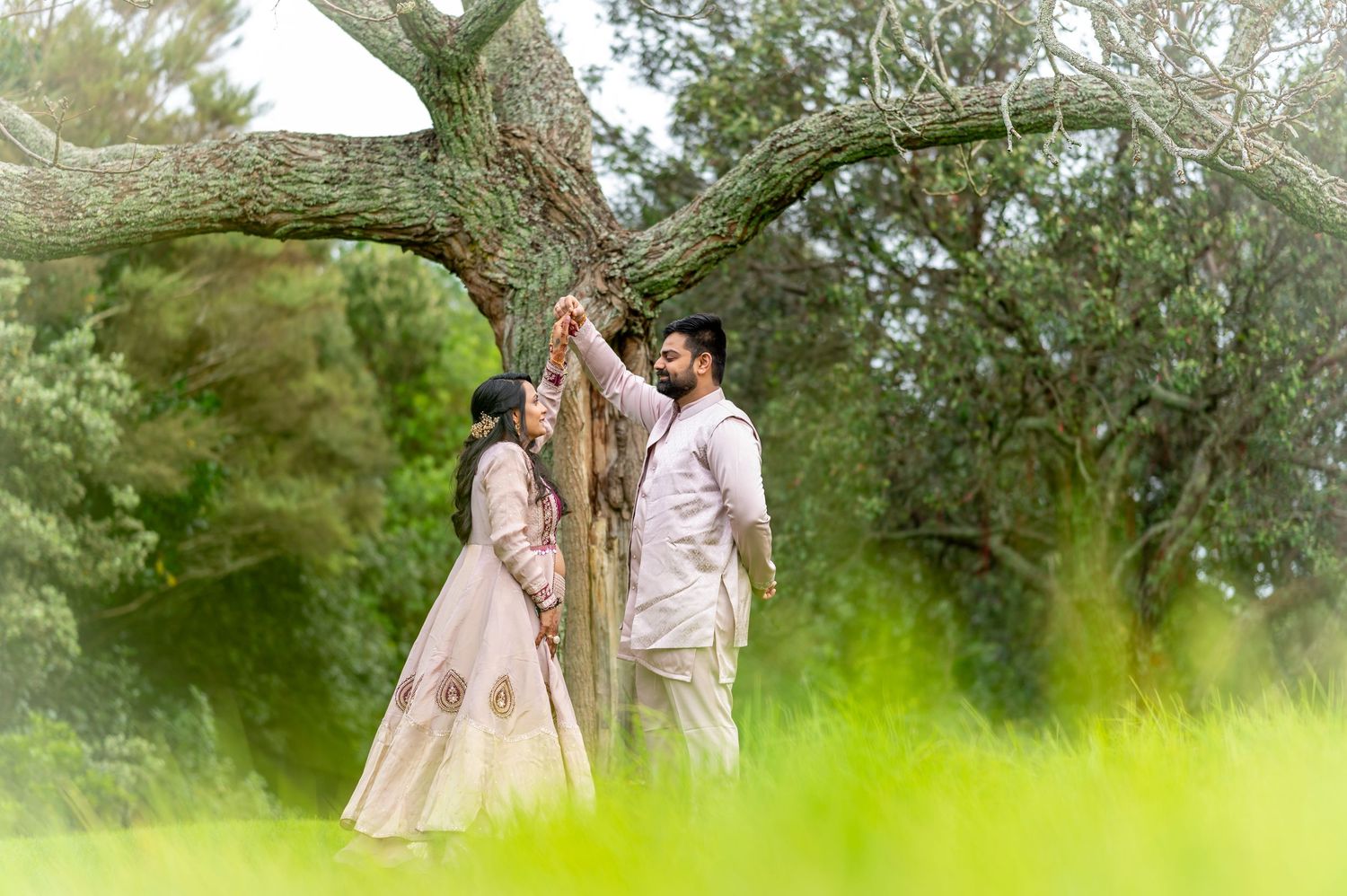 A romantic couple in traditional ethnic wedding attire stand beneath a sprawling oak tree in a lush green garden setting.