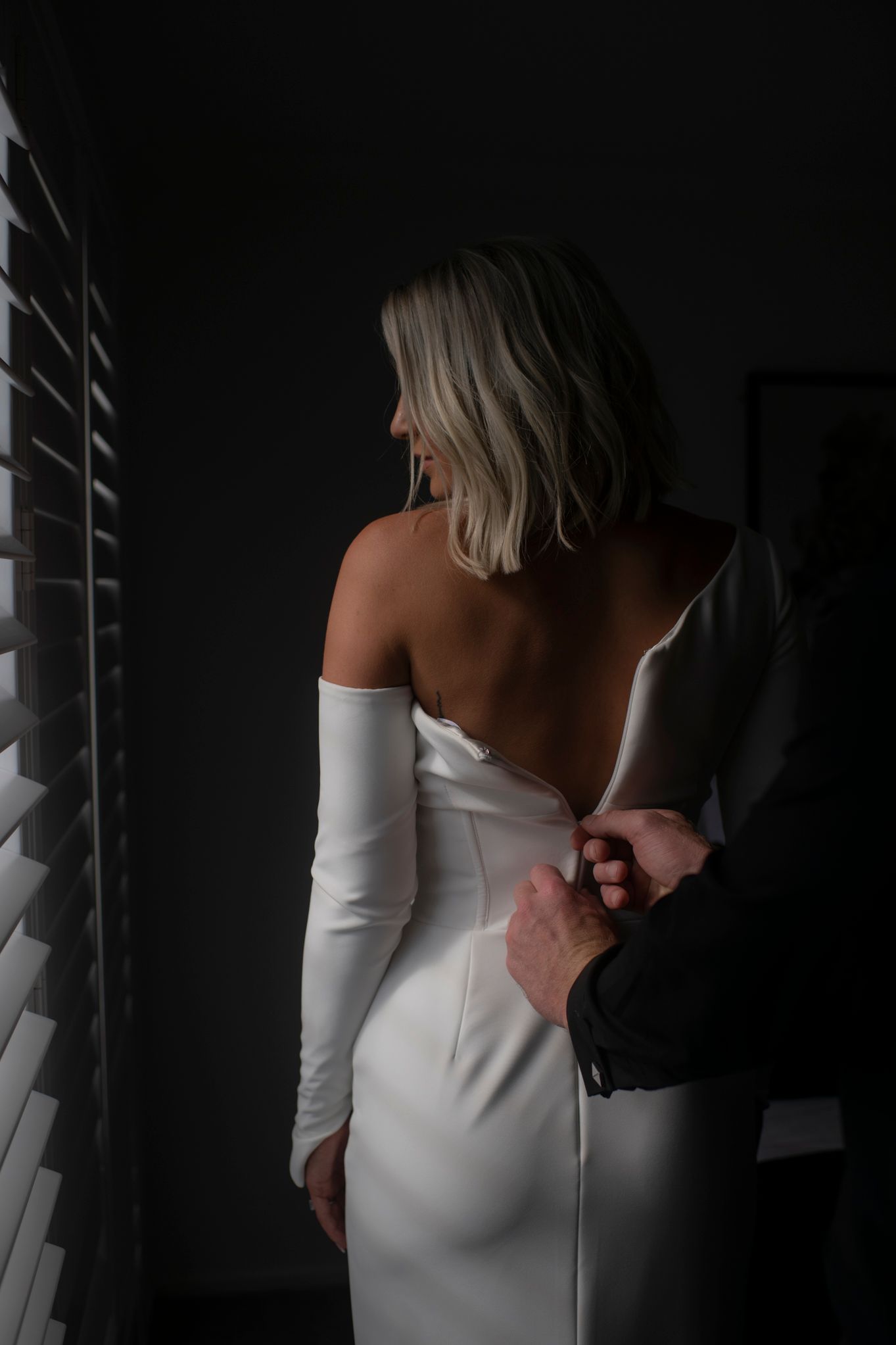 Woman in a sleek white off-shoulder dress poses elegantly against a dark background in an artistic photo series.