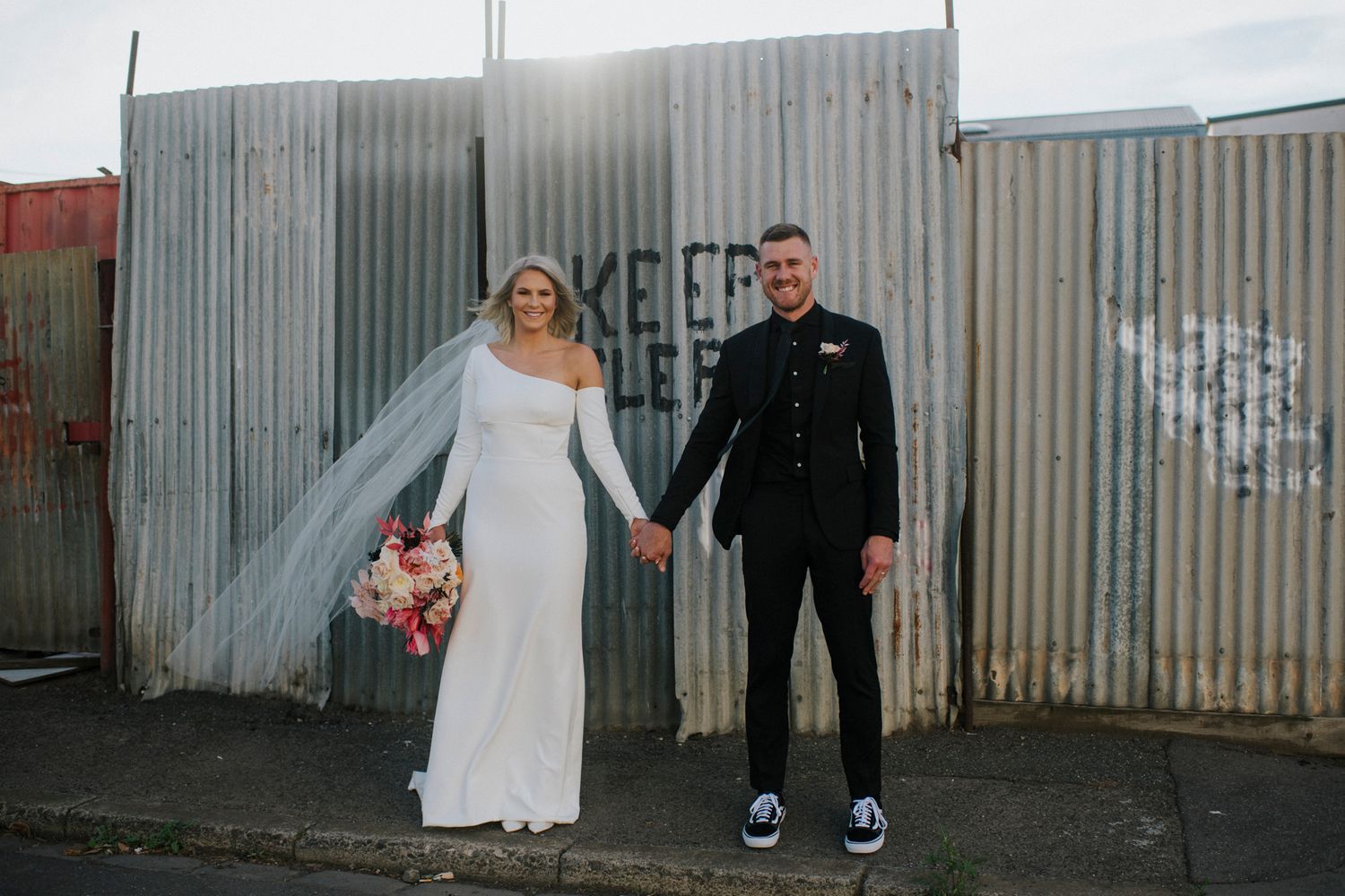A couple holds hands in front of corrugated metal wall, with flowing white dress and black formal attire.