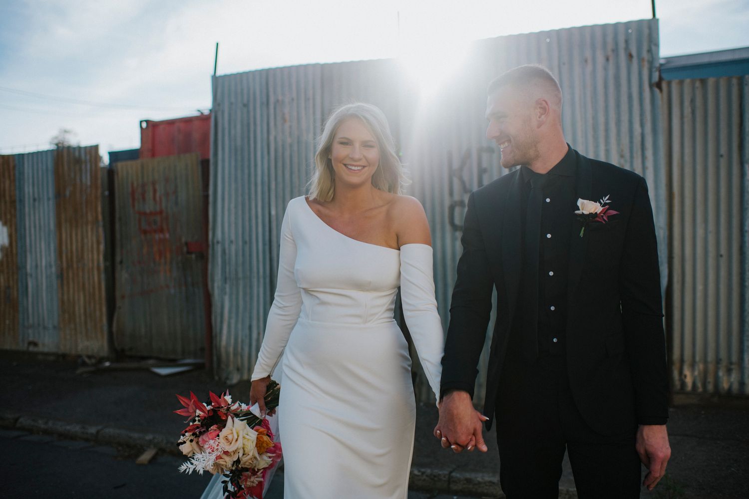 A wedding couple poses against an industrial backdrop with corrugated metal walls during their modern urban photoshoot.
