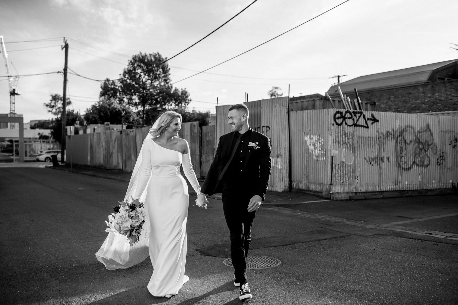 A couple in wedding attire walks down an urban street with graffiti walls and power lines in black and white.