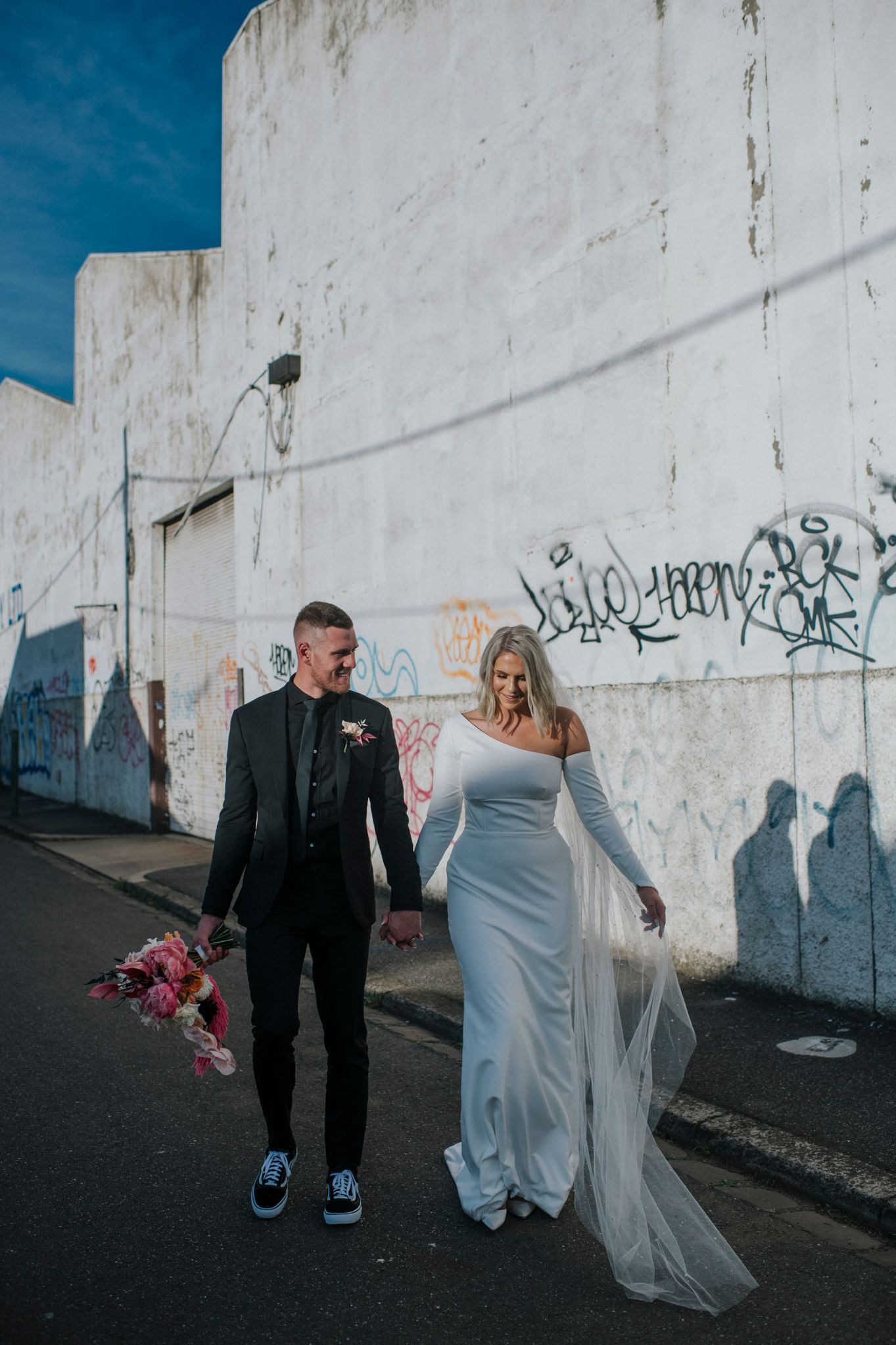 A couple walk hand in hand against a white wall with graffiti, dressed in formal wedding attire and blue sneakers.