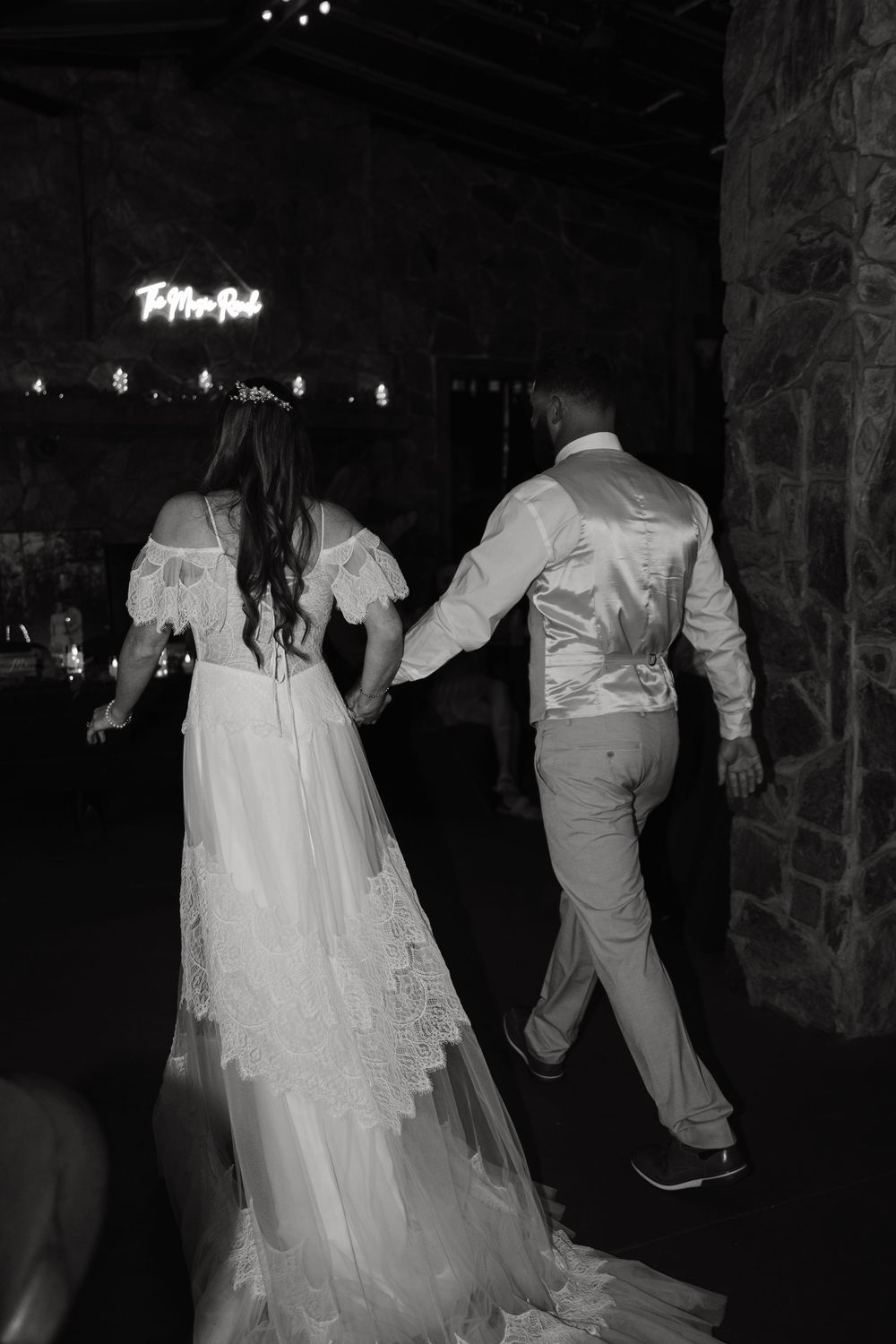 A bride and groom walk together in black and white, with her flowing lace gown trailing behind them.