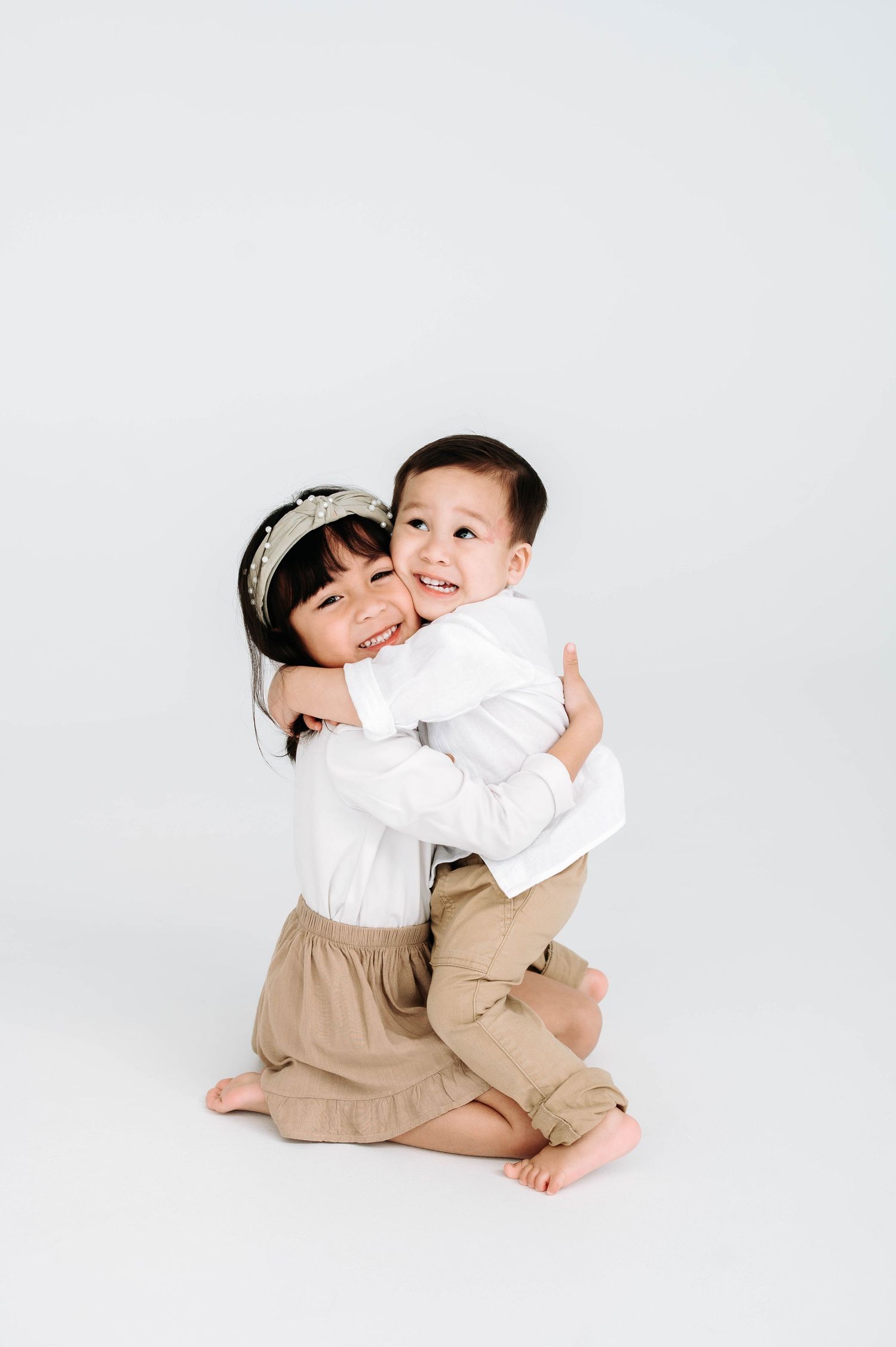 Two children in coordinated white and beige outfits share a sweet embrace on white background.