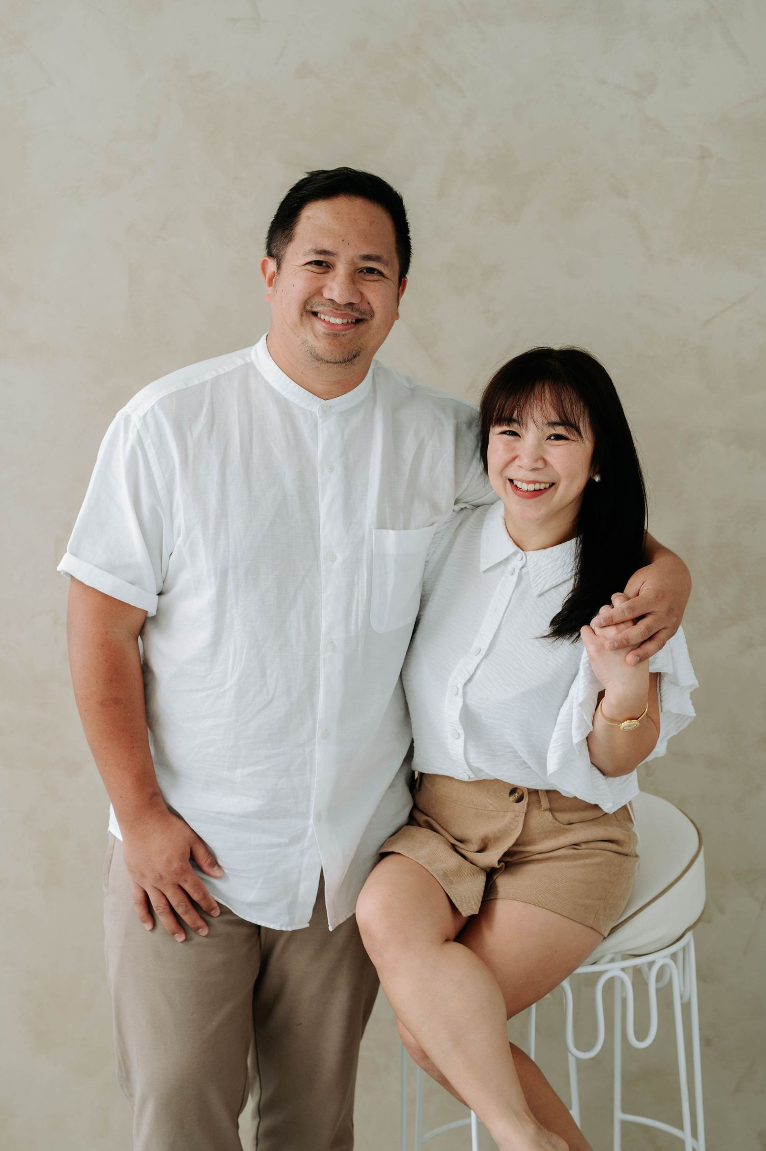 A couple wearing coordinated white shirts and khaki bottoms pose together against a light beige wall.