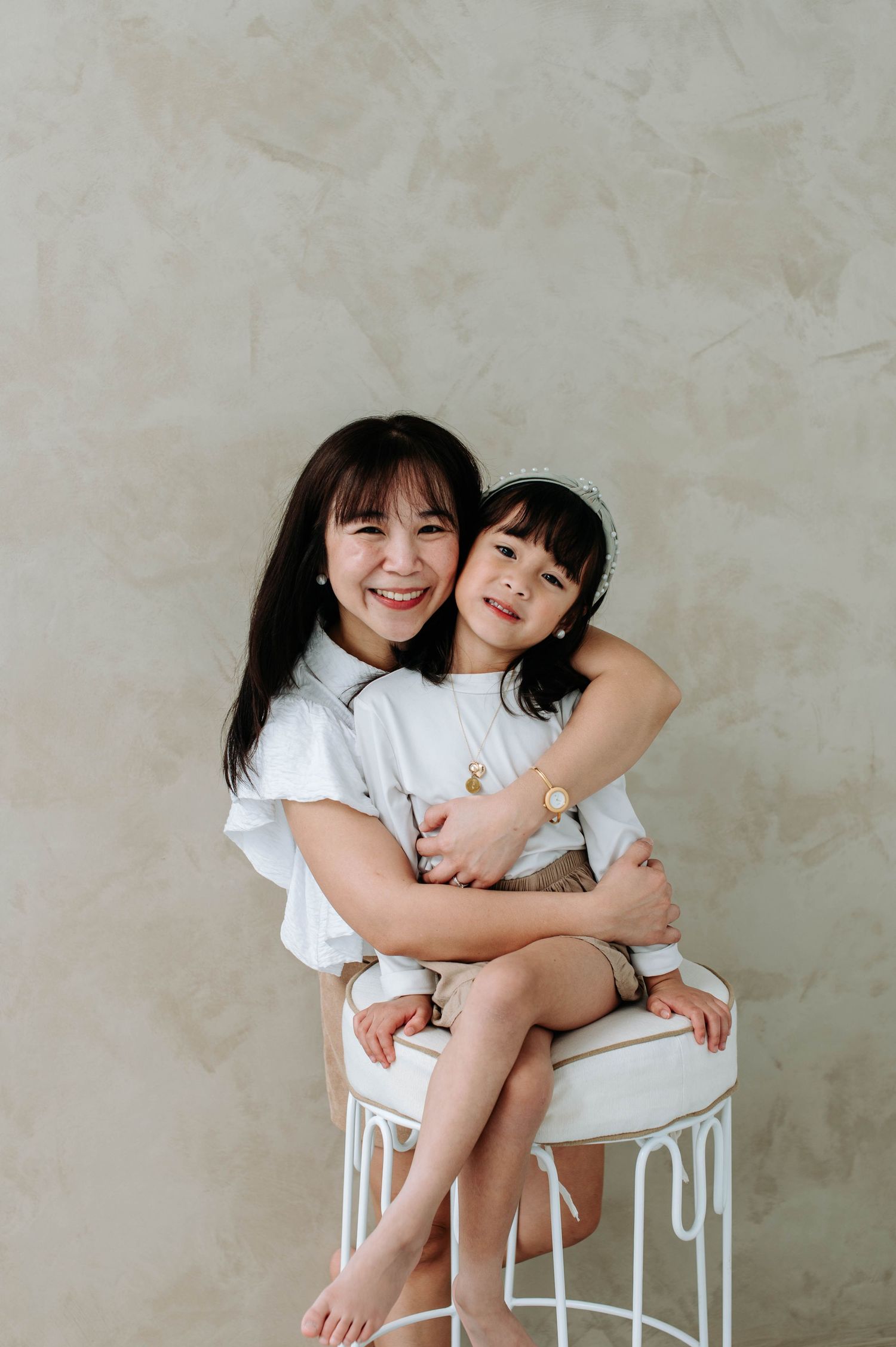 Two people share a warm embrace while sitting on a white stool against a neutral wall.