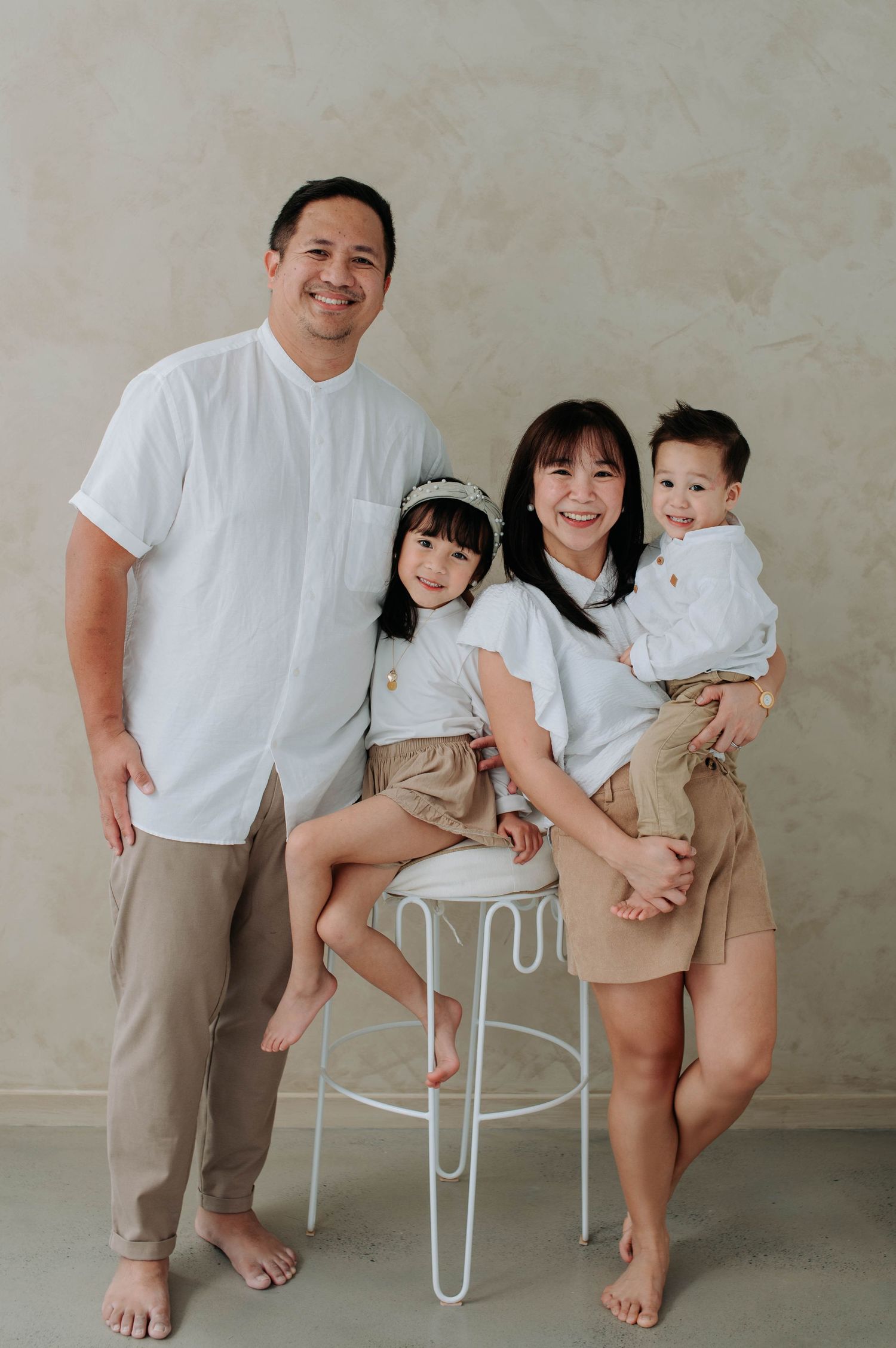 A family in matching white tops and khaki shorts poses playfully near a white chair against a neutral wall.