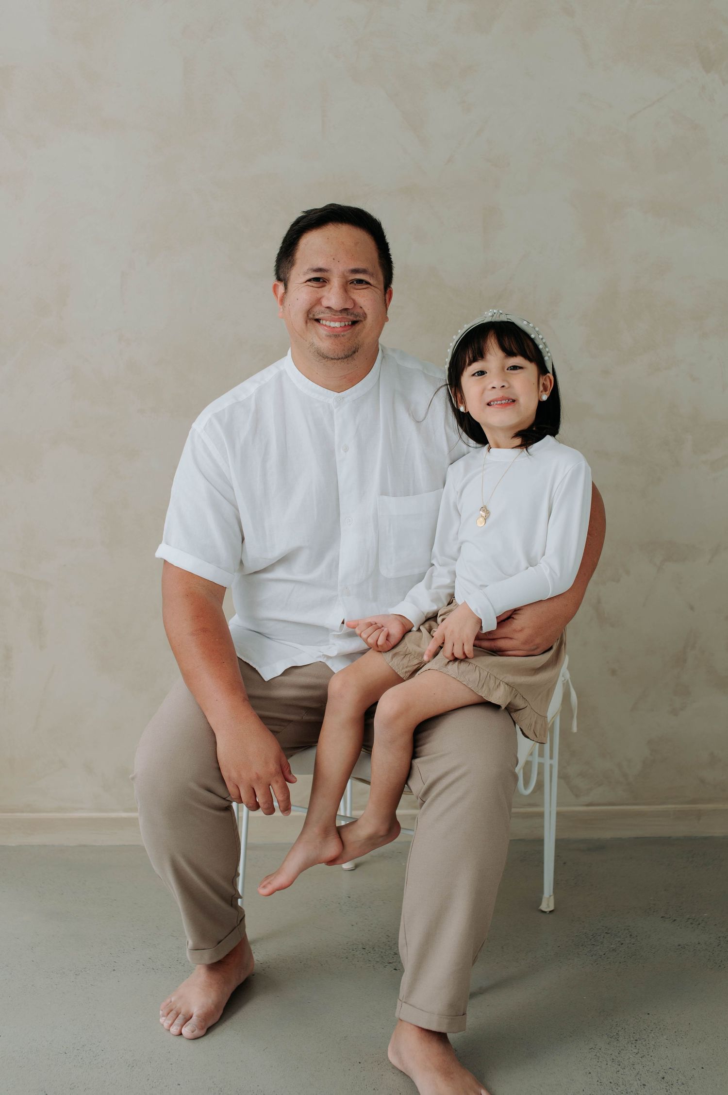 Family wearing coordinating white and beige outfits seated against neutral wall.