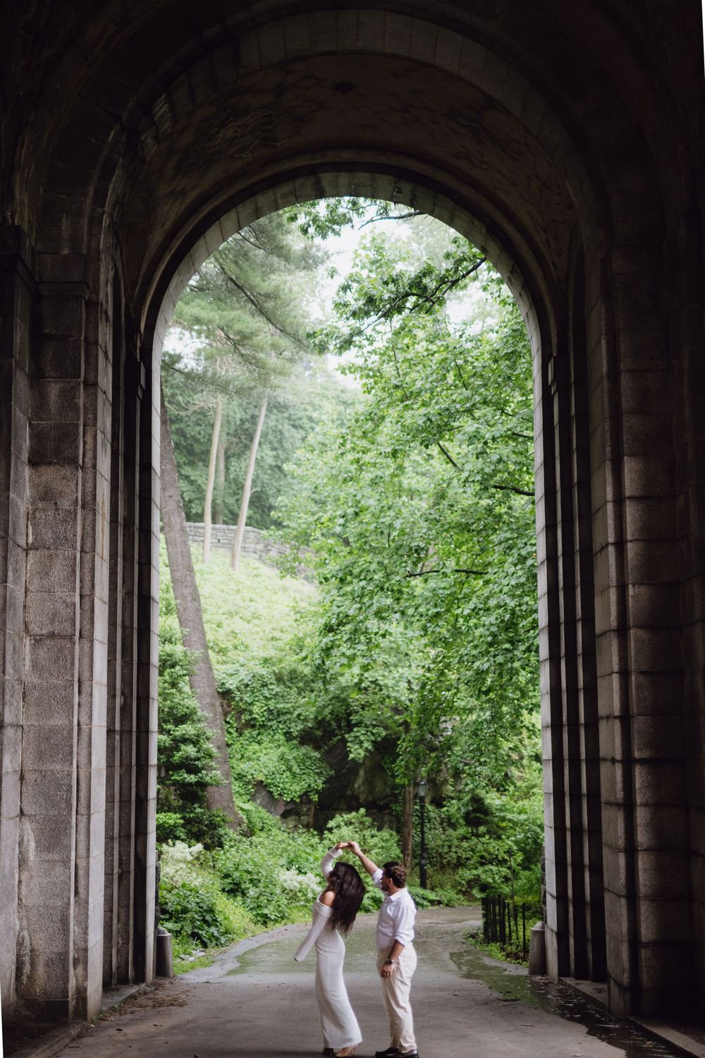 Two people in light colored clothing stand beneath a stone archway opening to a lush green forest path.