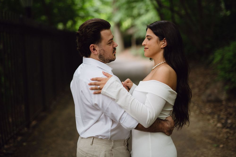 A romantic couple embraces on a tree-lined path, the bride in an off-shoulder white dress and groom in white shirt.