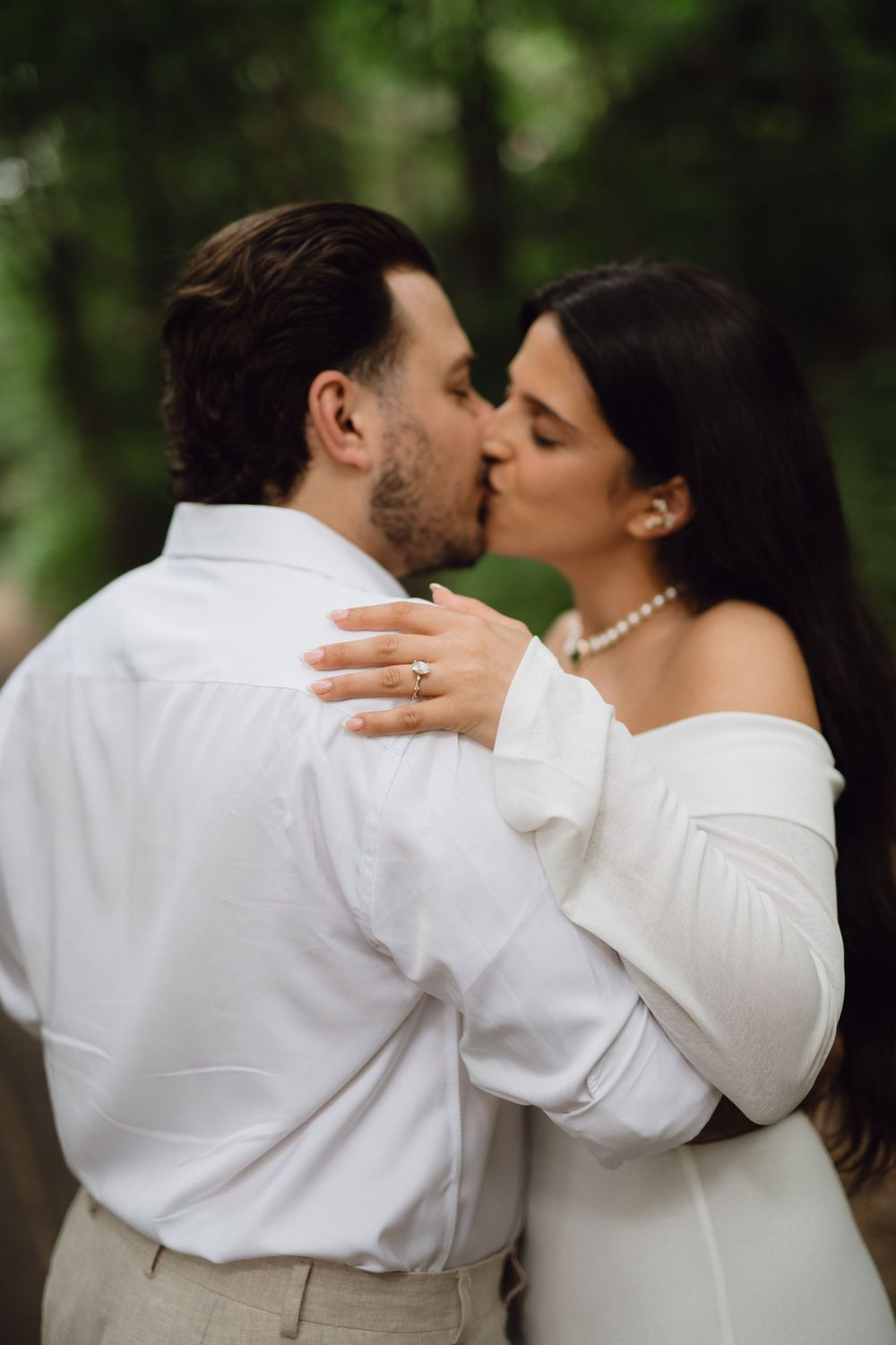 A tender romantic kiss between a couple in white attire against a blurred forest background.