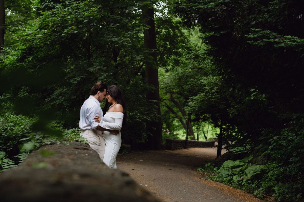 A couple shares an intimate moment while sitting on a stone wall surrounded by lush green forest scenery.