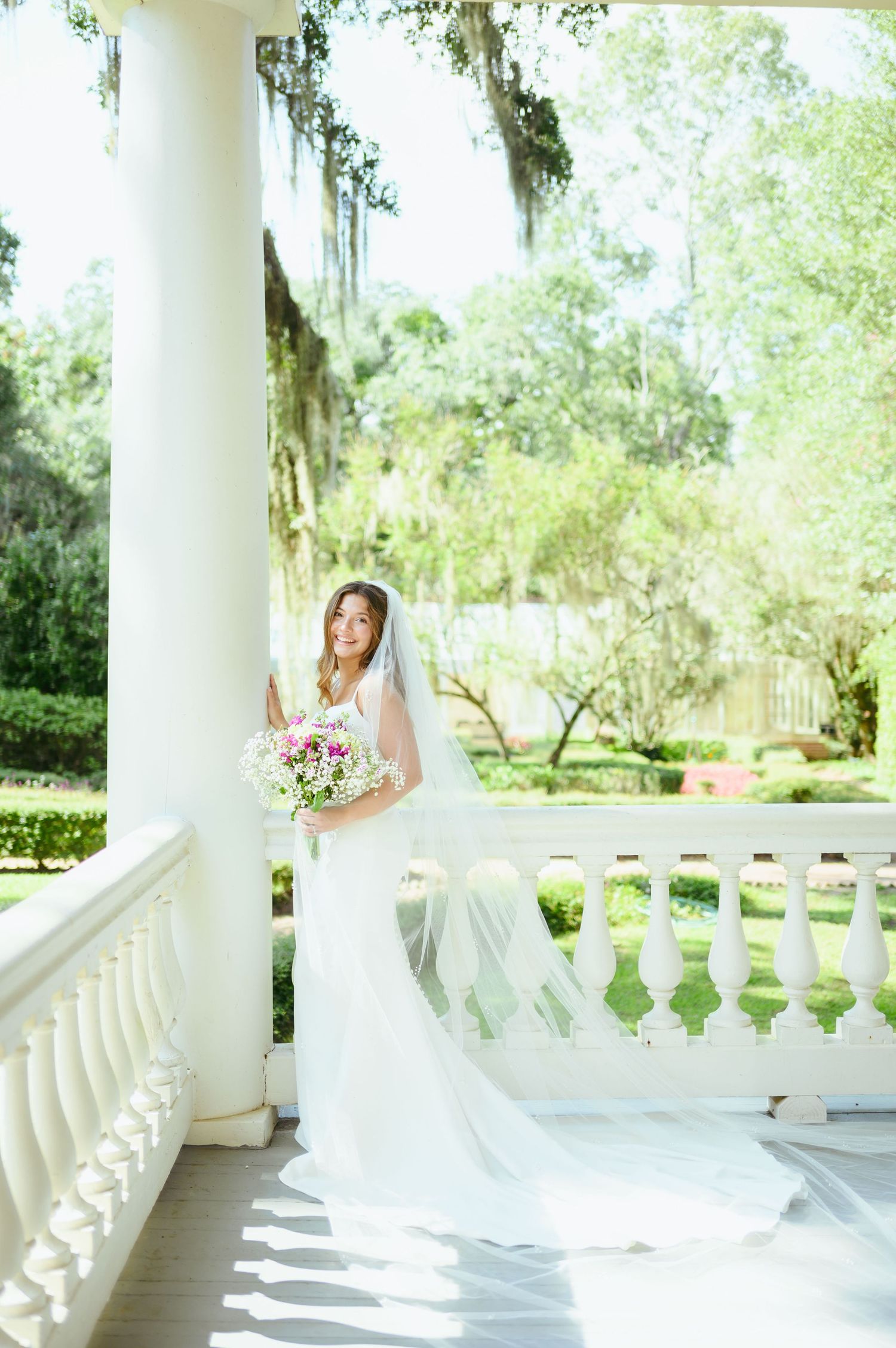 Elegant bride in white gown poses on classic white columned porch with bouquet.